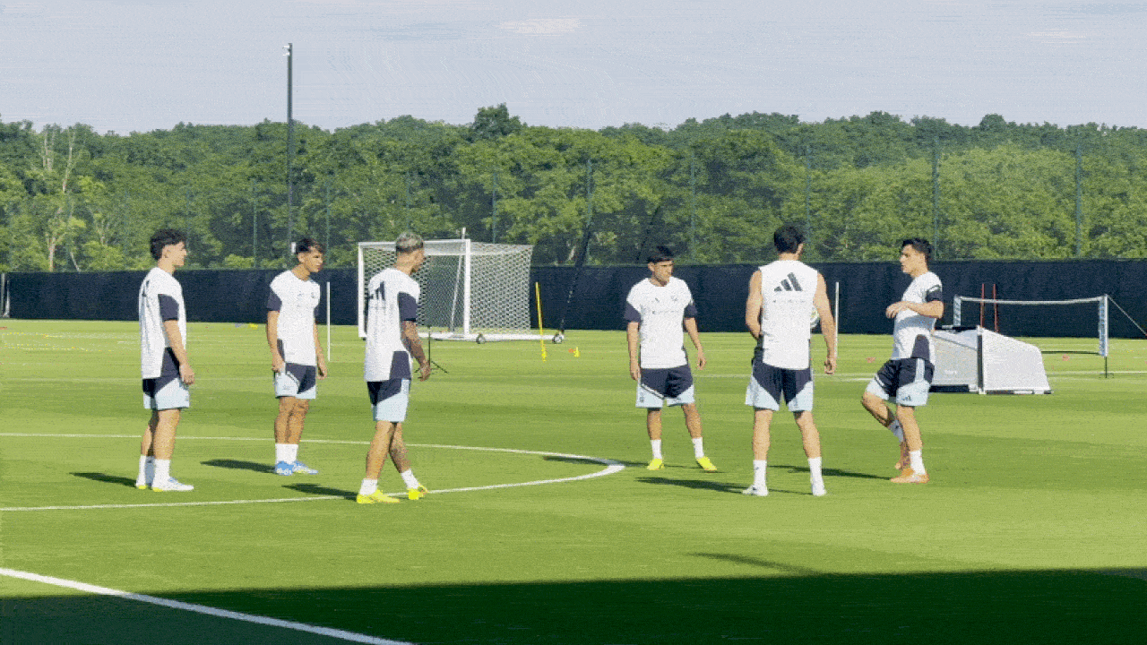 Six soccer players in white training kits stand on a bright green practice field near a goal, with cones and a netted barrier in the background, under a clear sky.