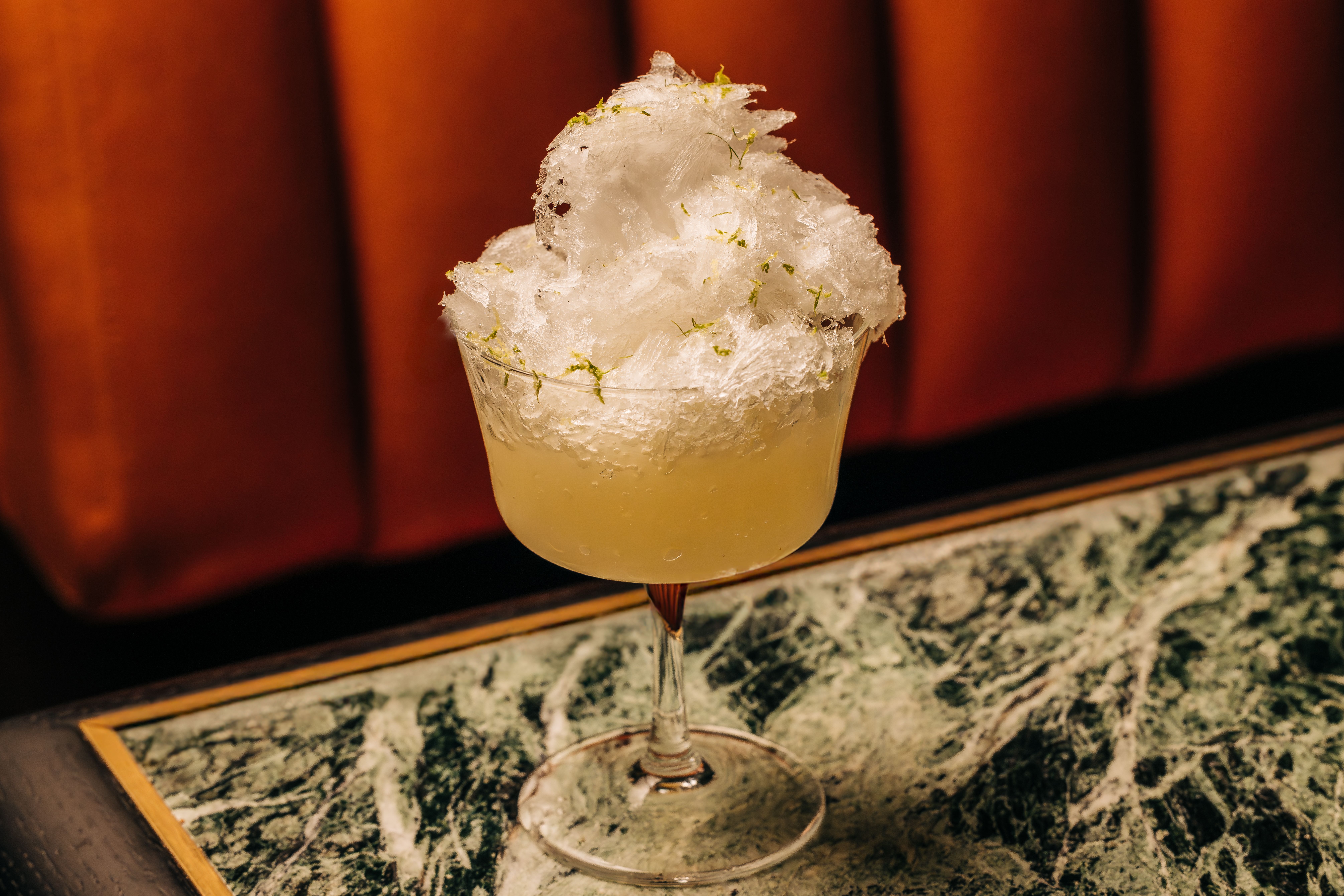 Clear cocktail glass filled with a pale yellow drink topped with a mound of crushed ice and green herb garnish, set on a green marble table with an orange-brown cushioned backdrop.