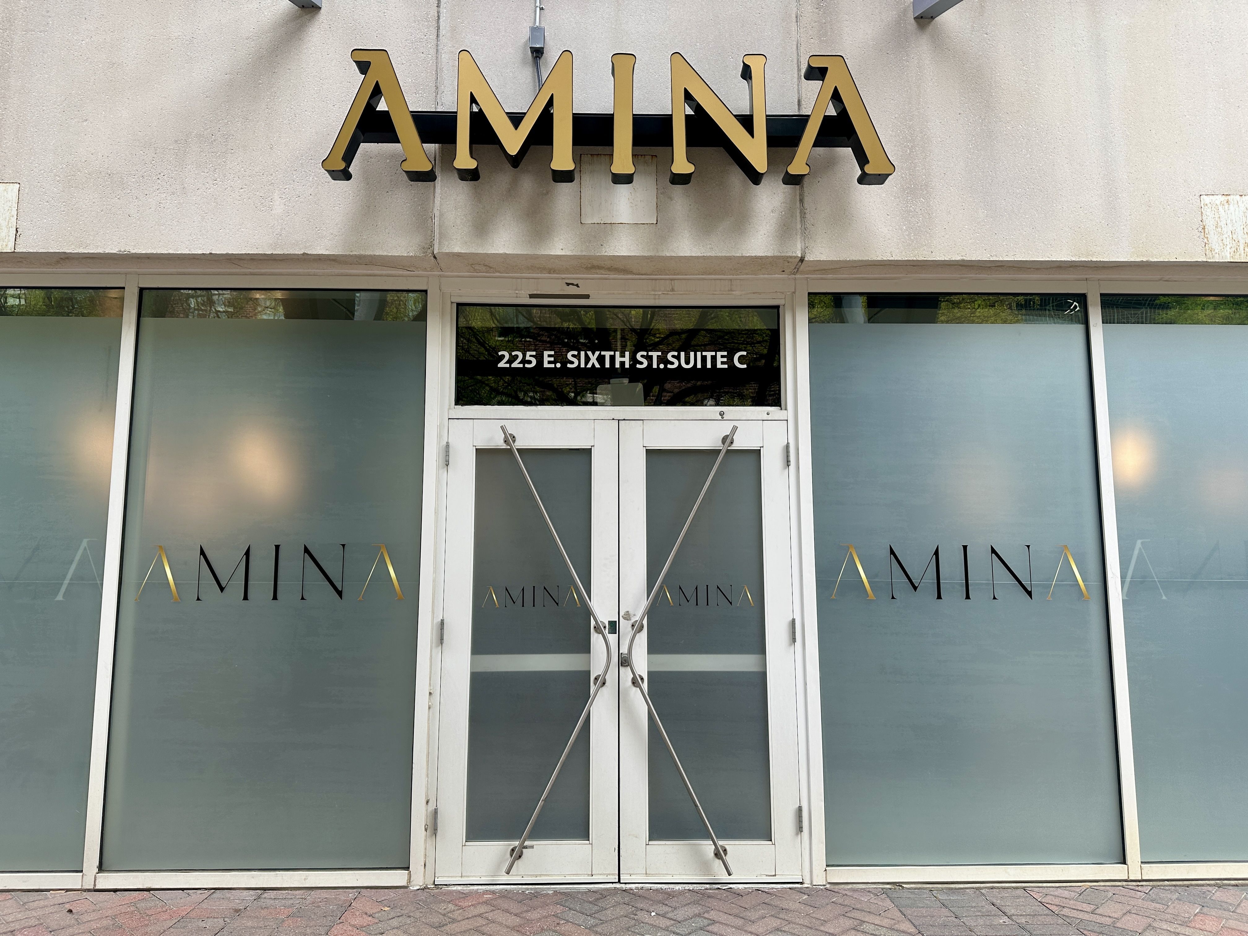 Beige concrete storefront with a large gold AMINA sign above white double doors. Frosted side windows show AMINA logos; address 225 E. SIXTH ST. SUITE C sits above the entry; doors have metal X-braces.