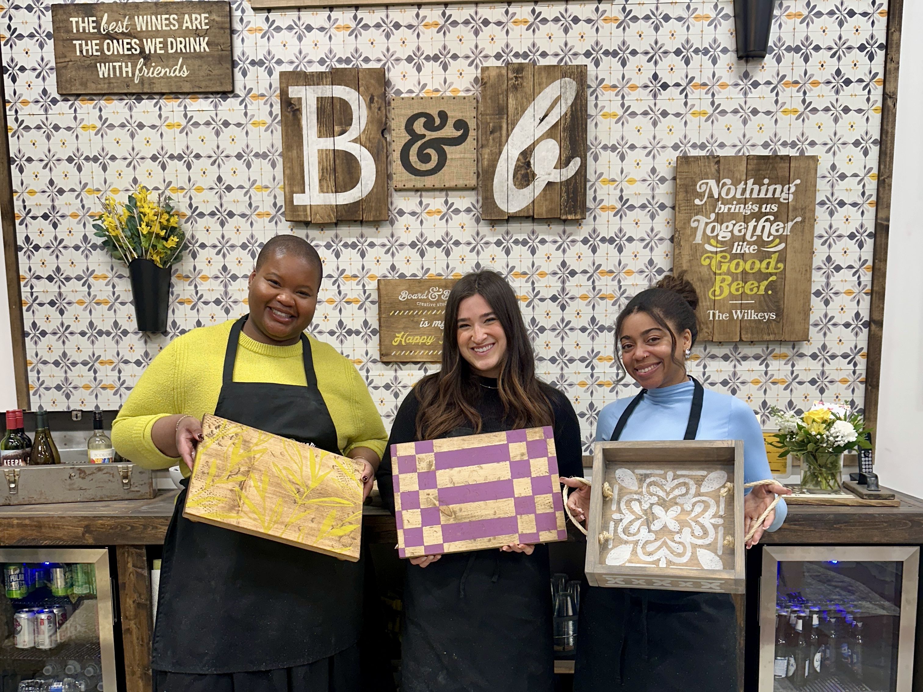 Board & Brush. Three smiling women in aprons pose behind a wooden bar, each holding a decorated board: yellow bamboo, purple-and-gold checker, and a white floral tile. A tiled wall with rustic signs behind them.