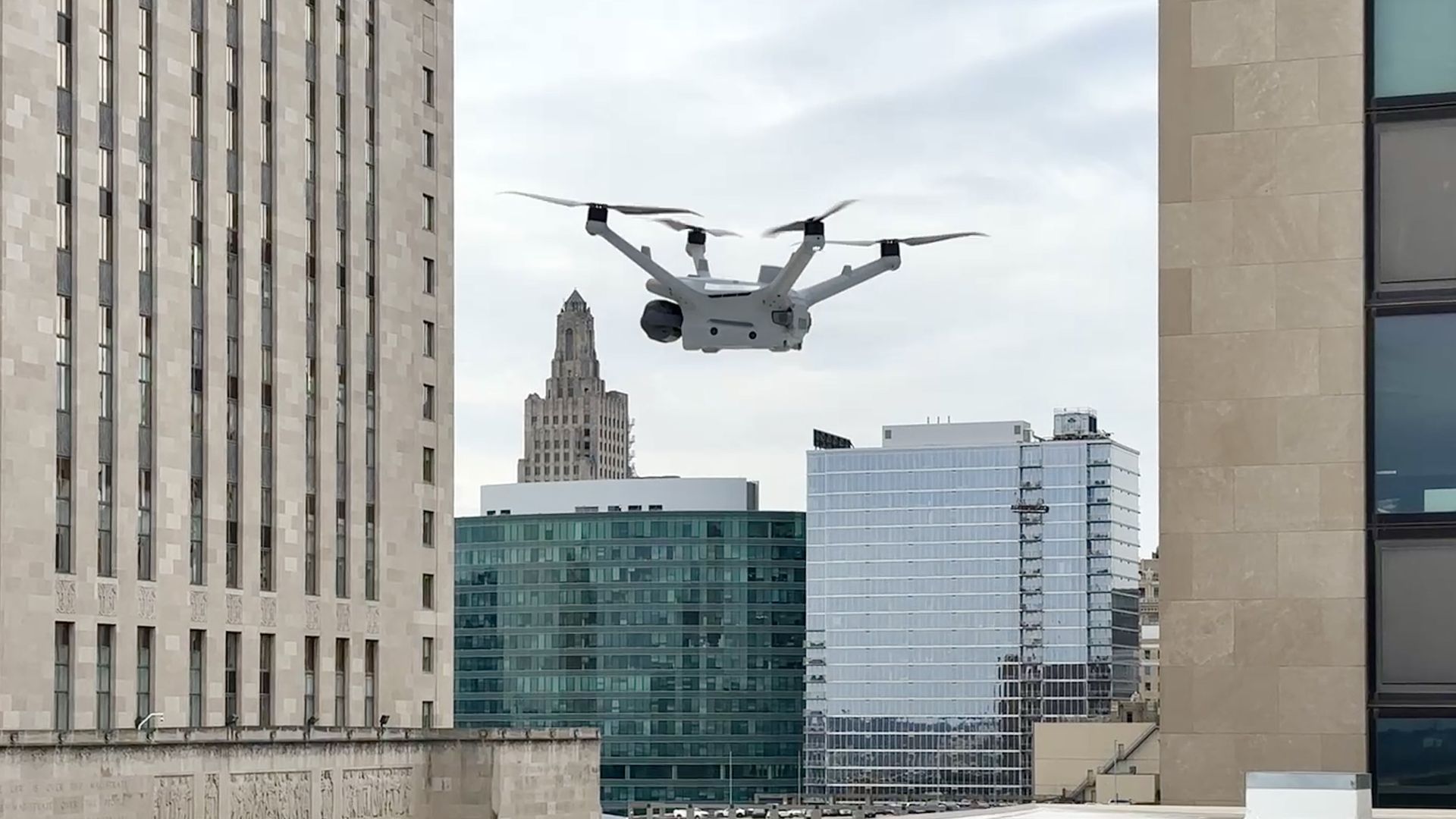 White quadcopter drone flying between tall modern and older buildings in a cityscape under a cloudy sky.