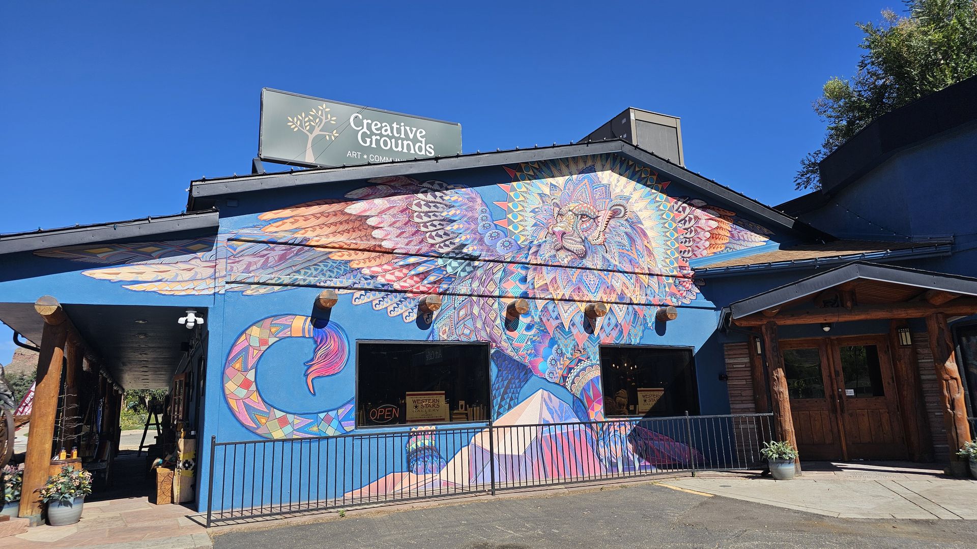 Blue building with a vibrant mural of colorful geometric wings and a central mandala; above, a sign reads "Creative Grounds" with "Art + Community" under a clear blue sky.