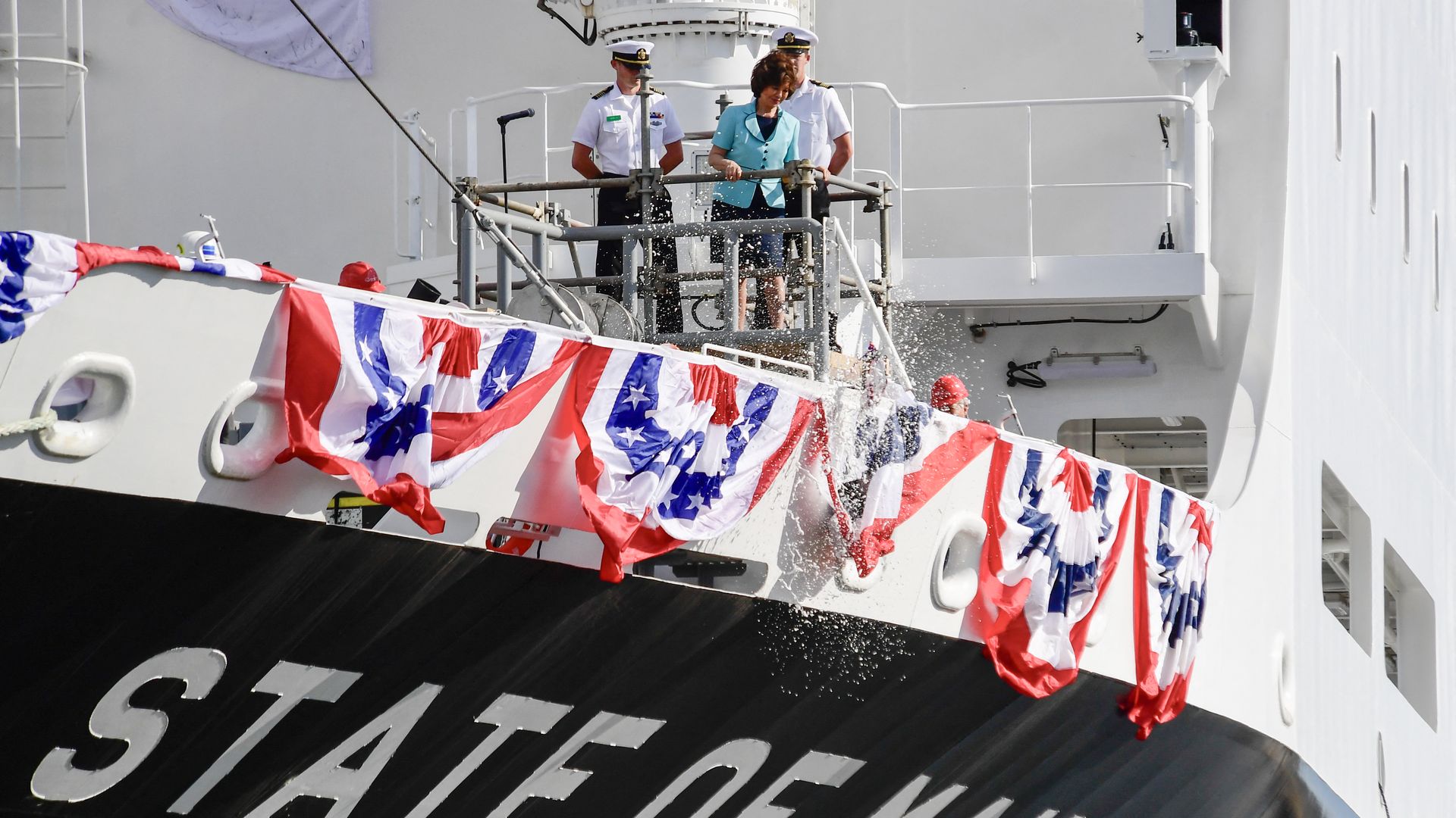 People standing on the deck of a ship called State of Maine, breaking bottles over the edge, draped with flag bunting. 