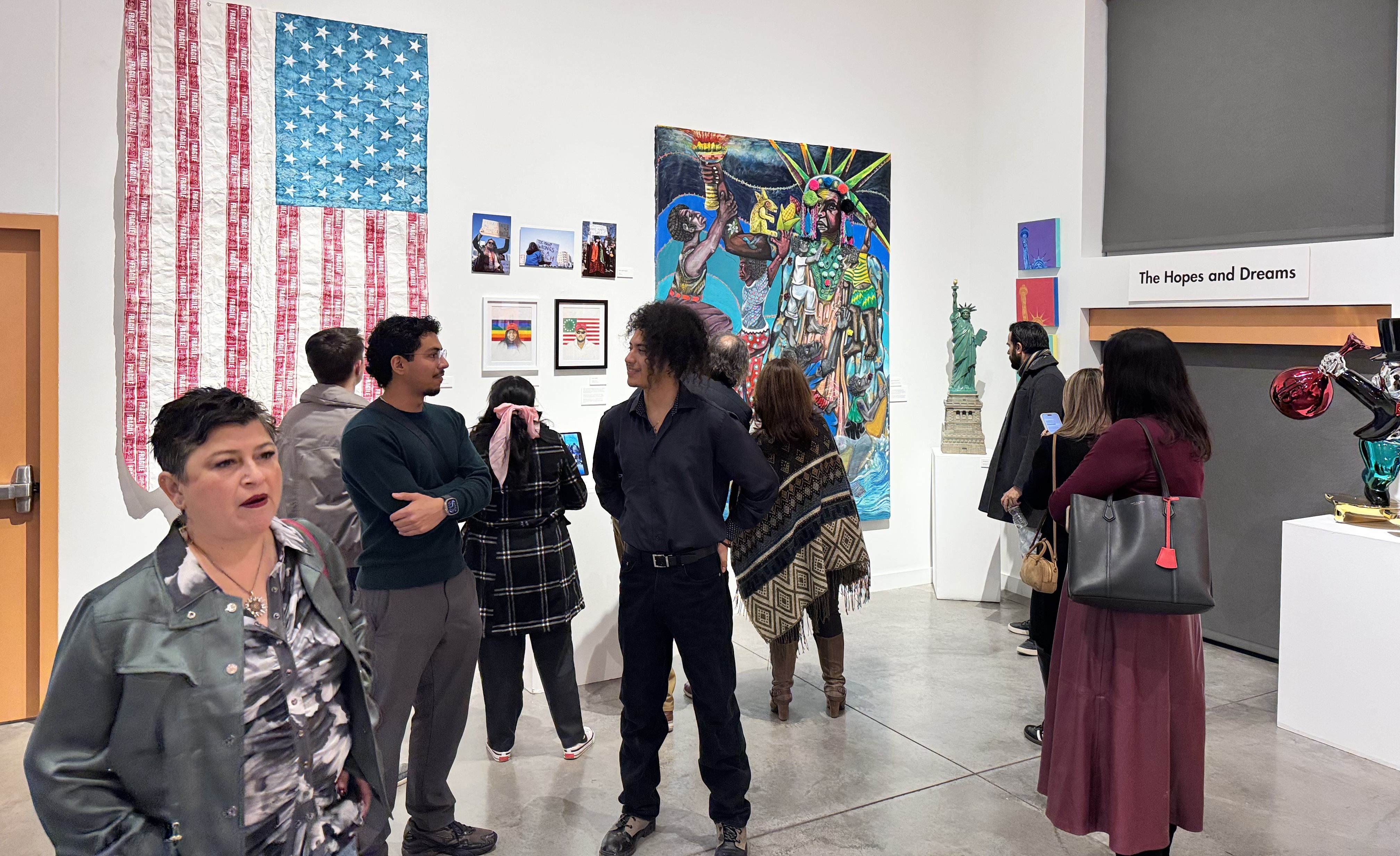 People viewing diverse artwork in a gallery, including an American flag with red text stripes, colorful paintings, and a Statue of Liberty sculpture under a sign saying "The Hopes and Dreams".