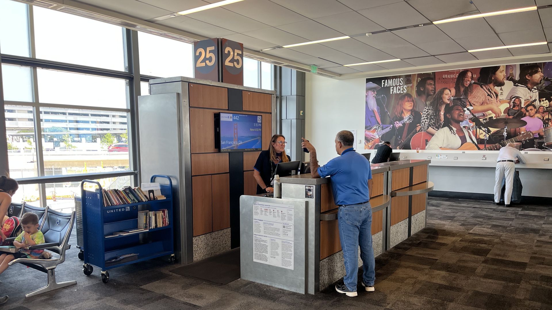 Airport check-in area with a wood-front desk and screen showing 'San Francisco, CA.' A staffer assists a traveler as large '25' signs hang from the ceiling; a mural of a live band spans the far wall.
