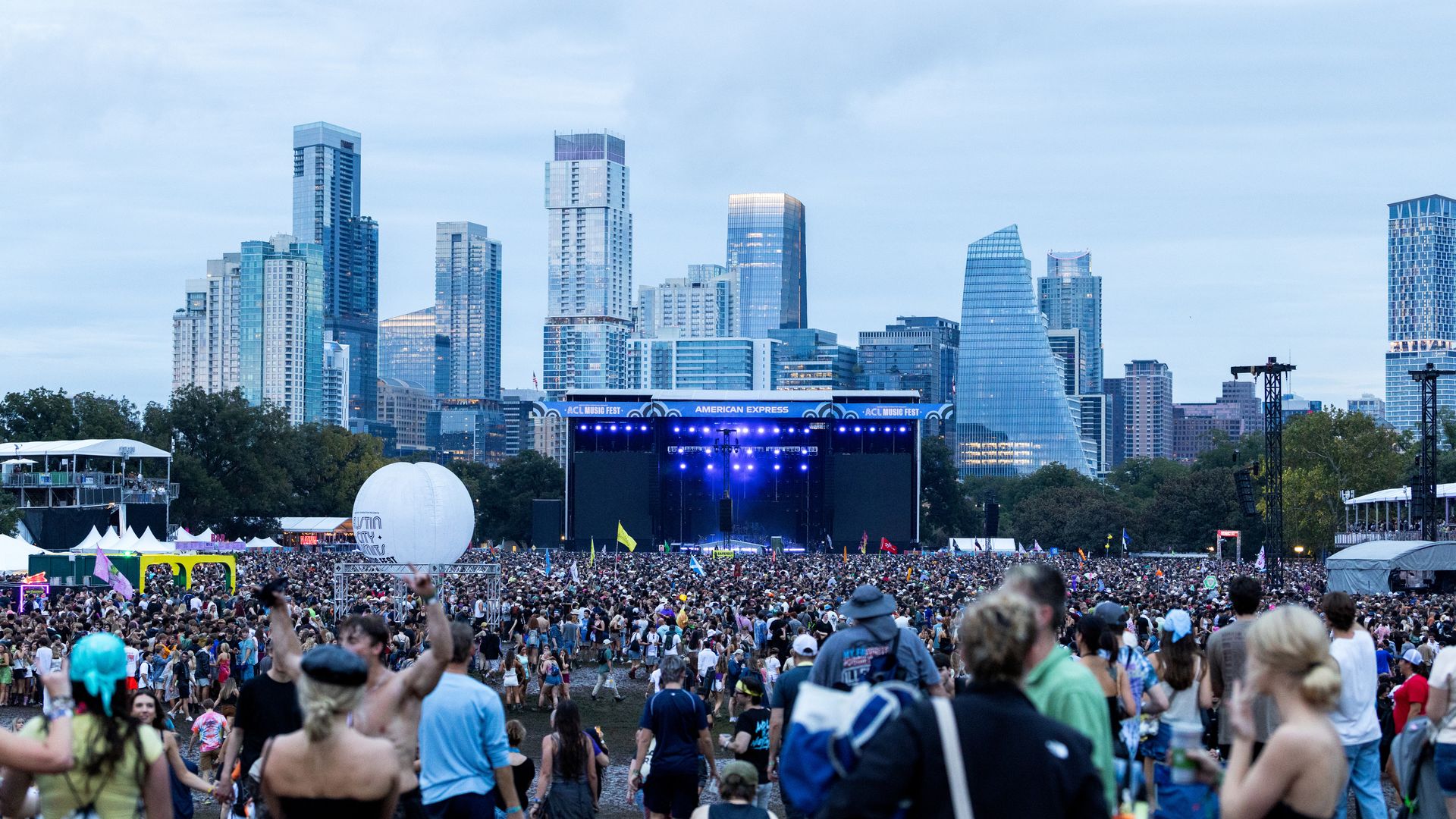 Large crowd at an outdoor concert in a city park, stage with blue lights, and a modern downtown skyline of tall glass buildings behind, under a cloudy sky.