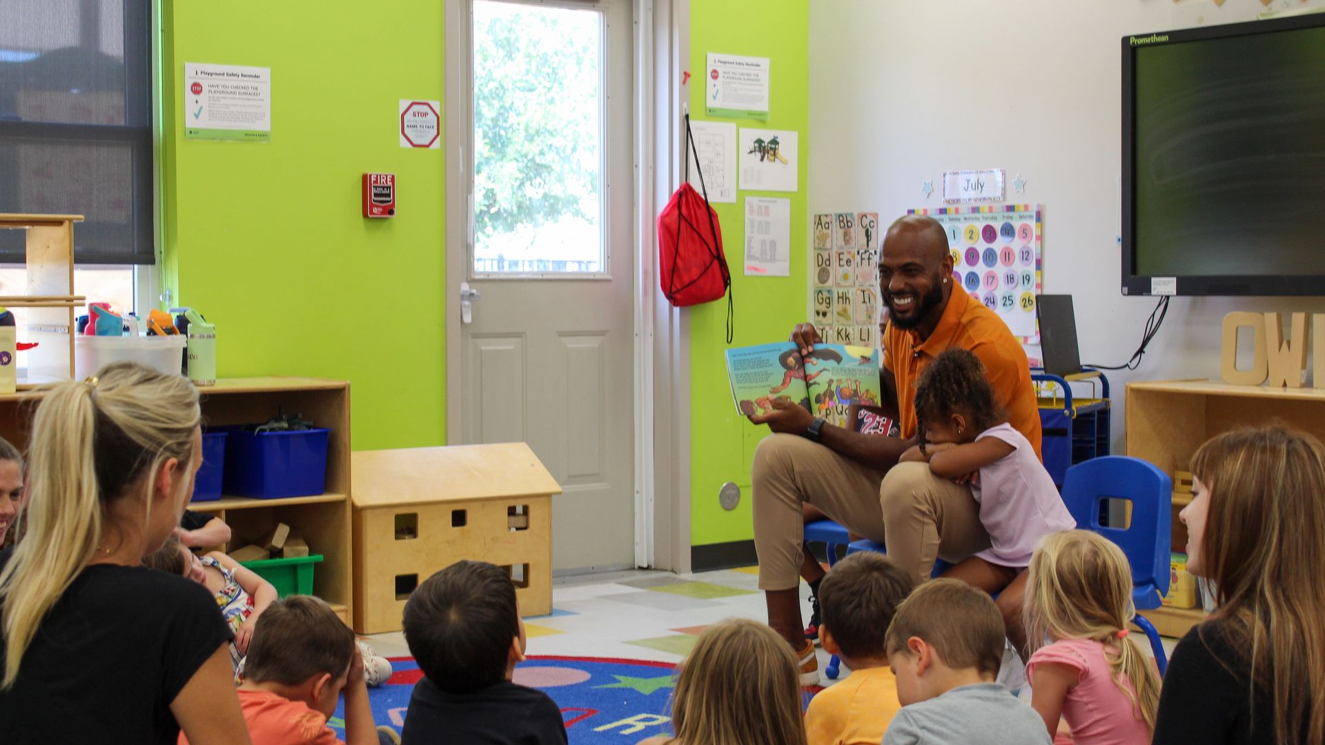 Derrick Johnson in an orange shirt reading a colorful children’s book to a group of young kids seated on a blue and multicolored rug in a bright classroom with green and white walls.