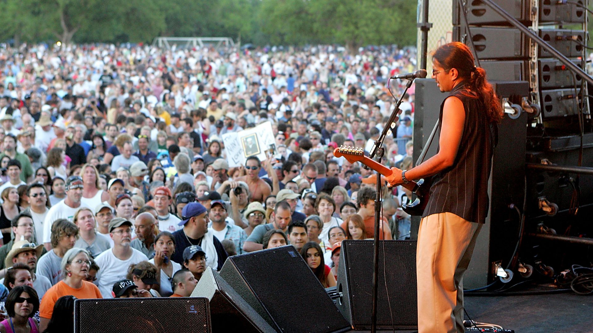 A guitarist performs on stage with a crowd of onlookers in the background.