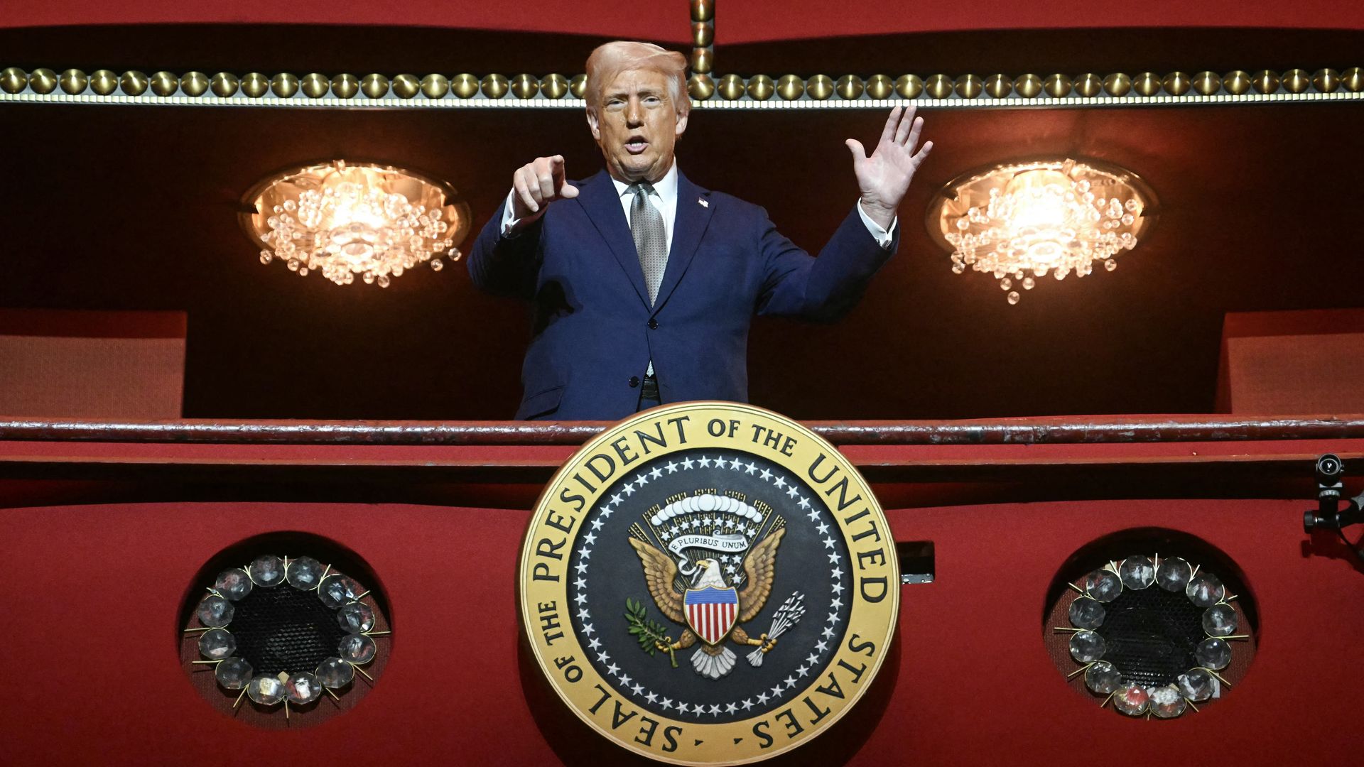 President Trump, wearing a blue suit and pointing at the camera while standing on a red balcony with the presidential seal on it.