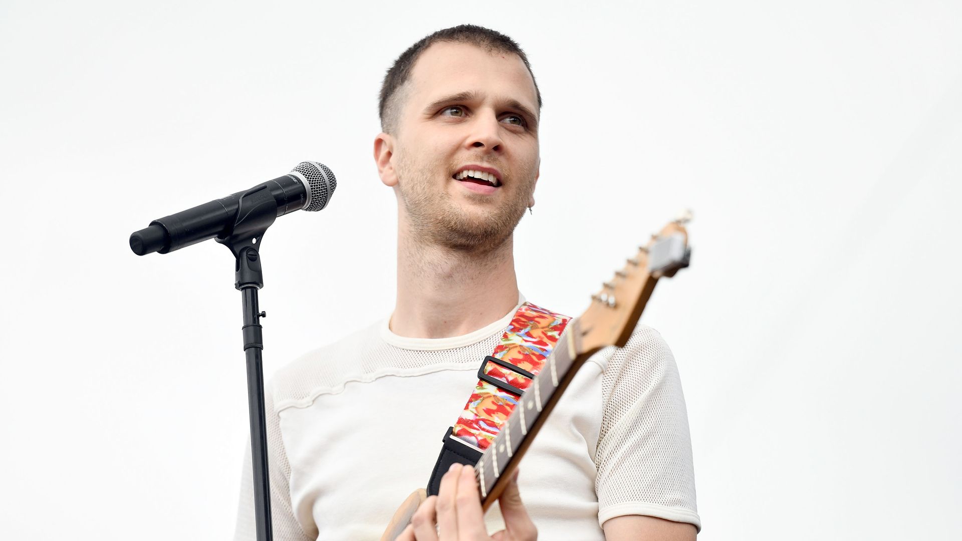 A man with short brown hair wearing a white T-shirt (singer JMSN) plays a guitar as he smiles from behind a microphone during a music festival.