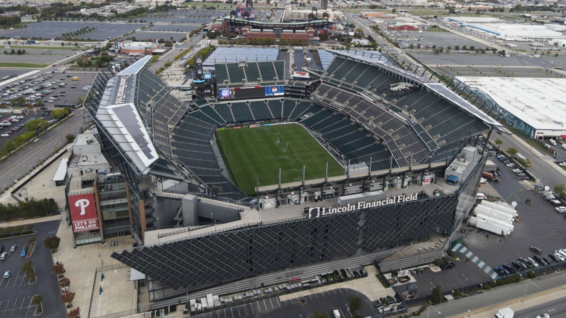 Aerial view of Lincoln Financial Field stadium with green field, mostly empty dark green seats, and surrounding cityscape under cloudy sky.