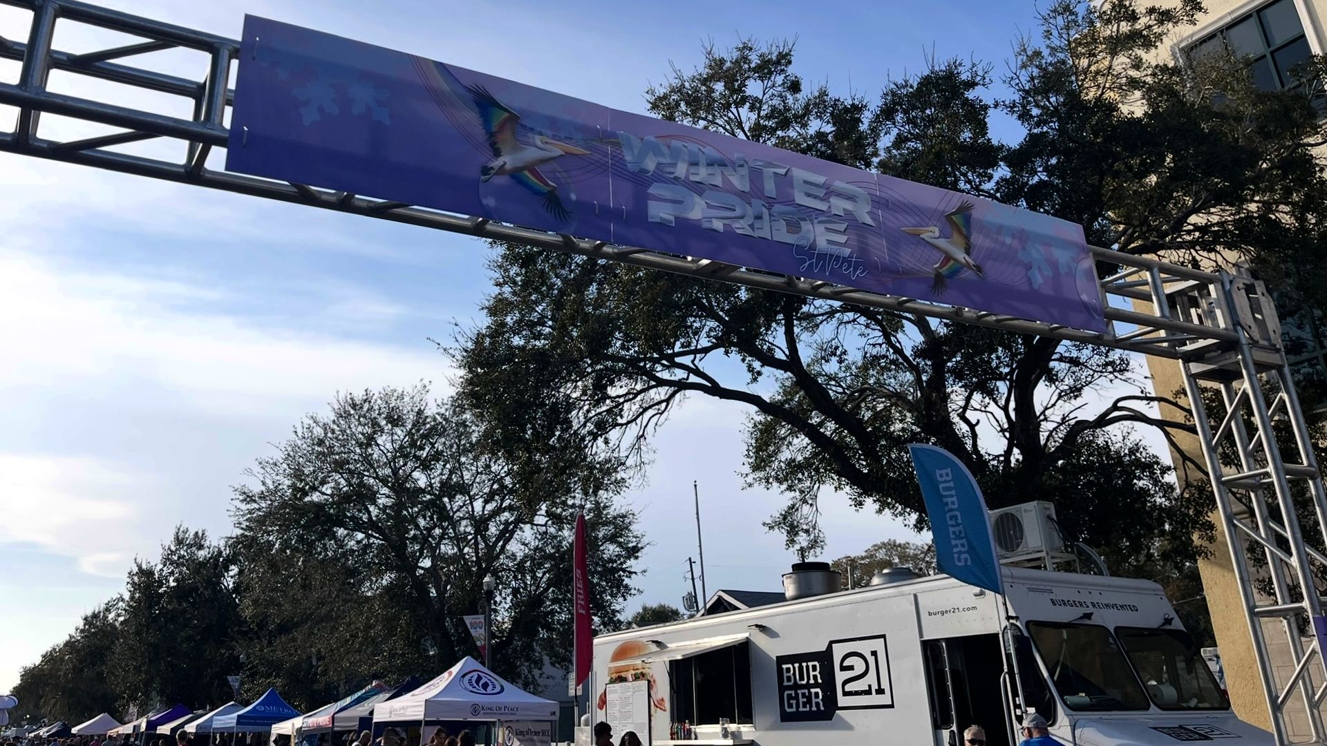 Outdoor event with a purple "Winter Pride St. Pete" banner featuring pelicans, food trucks including Burger 21, tents, and people under a clear sky with trees in the background.