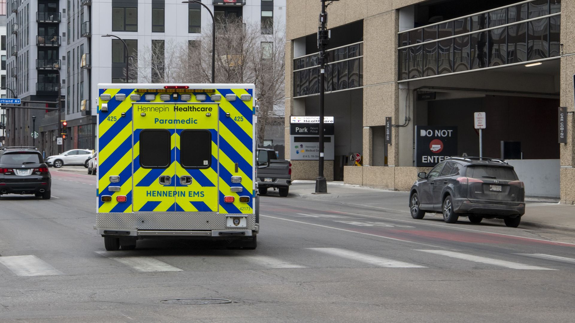Rear view of a Hennepin Healthcare paramedic ambulance with yellow and blue stripes stopped at a red traffic light on a city street with tall buildings and parked cars.