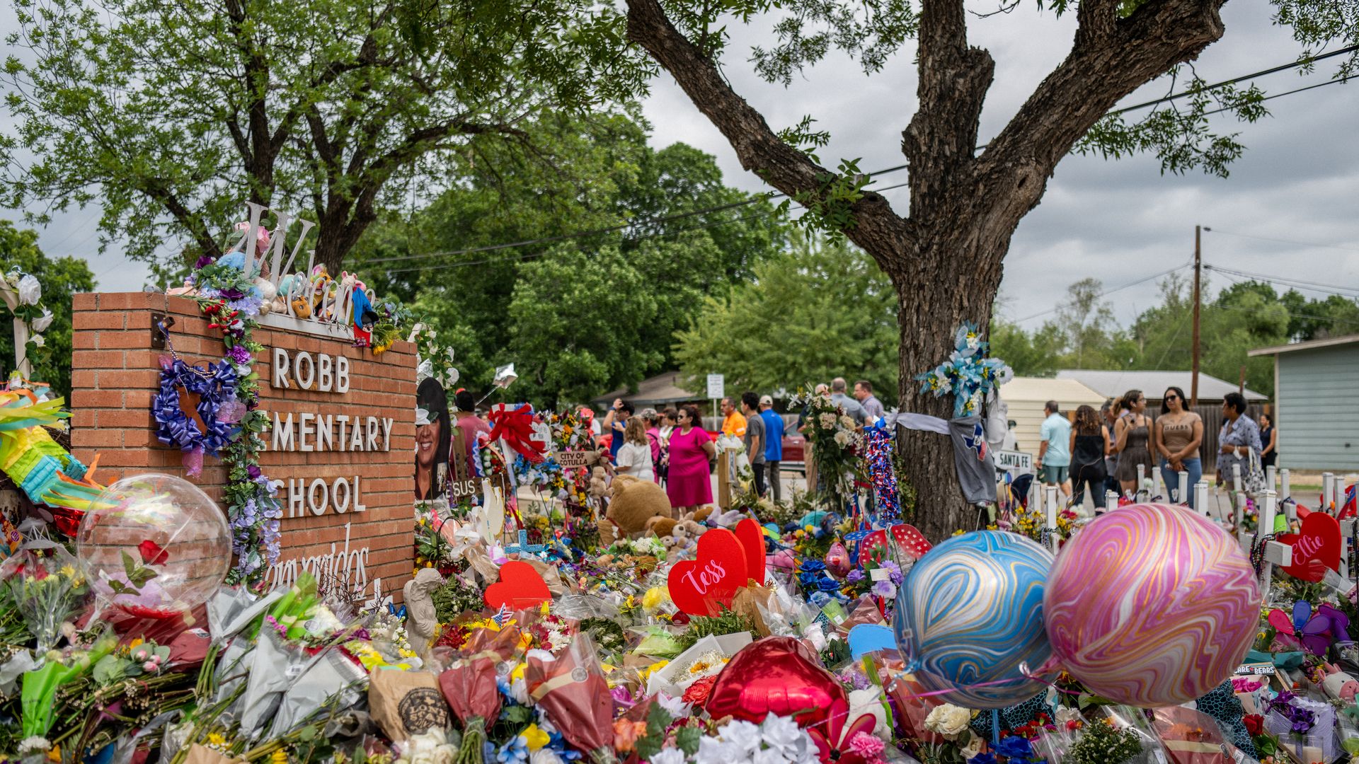 Photo of flowers, balloons, gifts and other memorabilia crowded around a brick sign that says "Robb Elementary School"