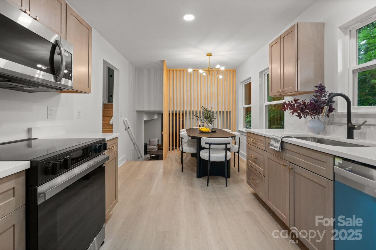 Modern kitchen with light wood cabinets, stainless steel appliances, black faucet, white countertops, dining table with chairs, decorative plants, and wooden slat room divider.