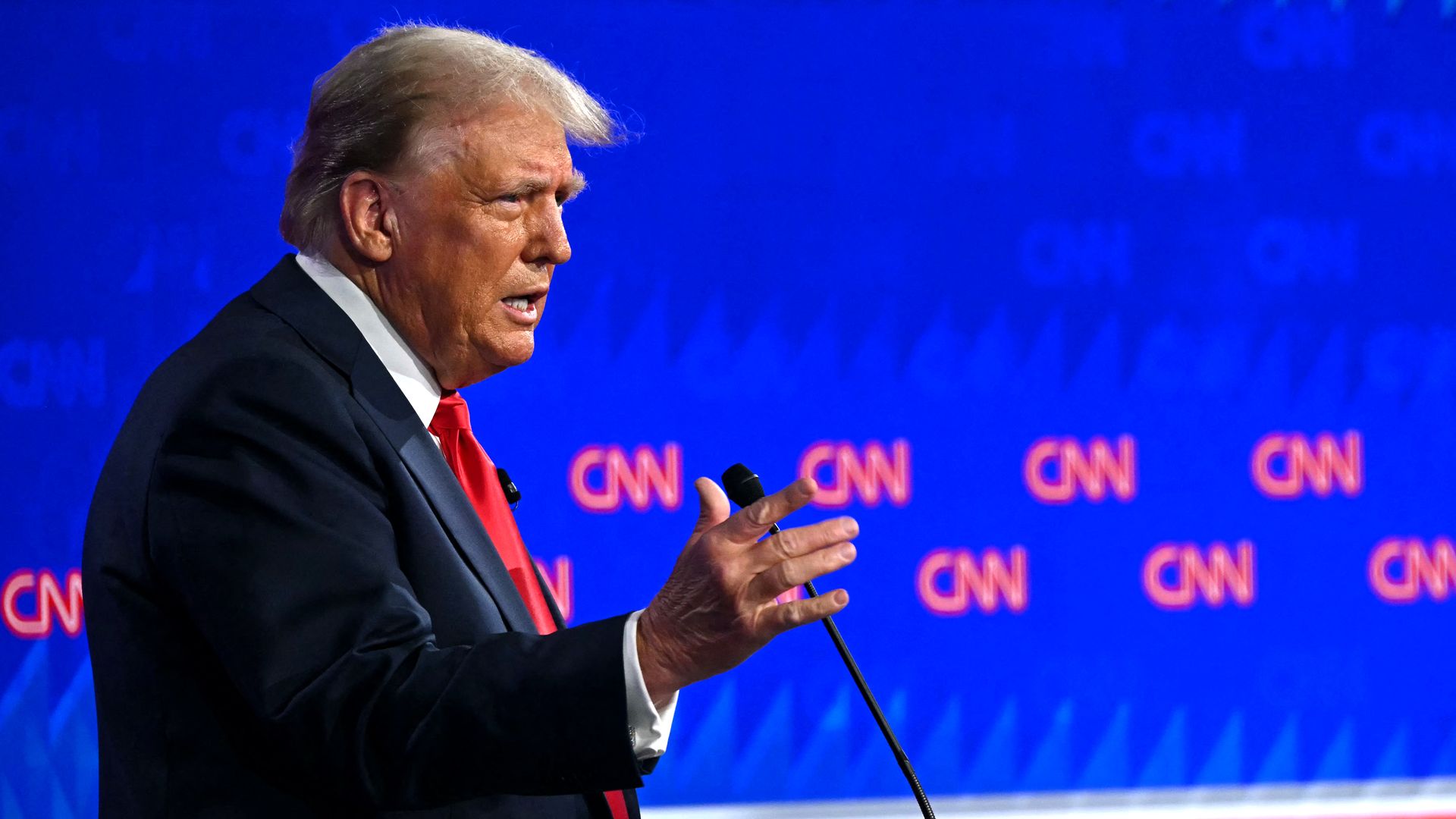 Former President Trump, wearing a blue suit, white shirt and red tie, speaking into a microphone in front of a CNN backdrop.