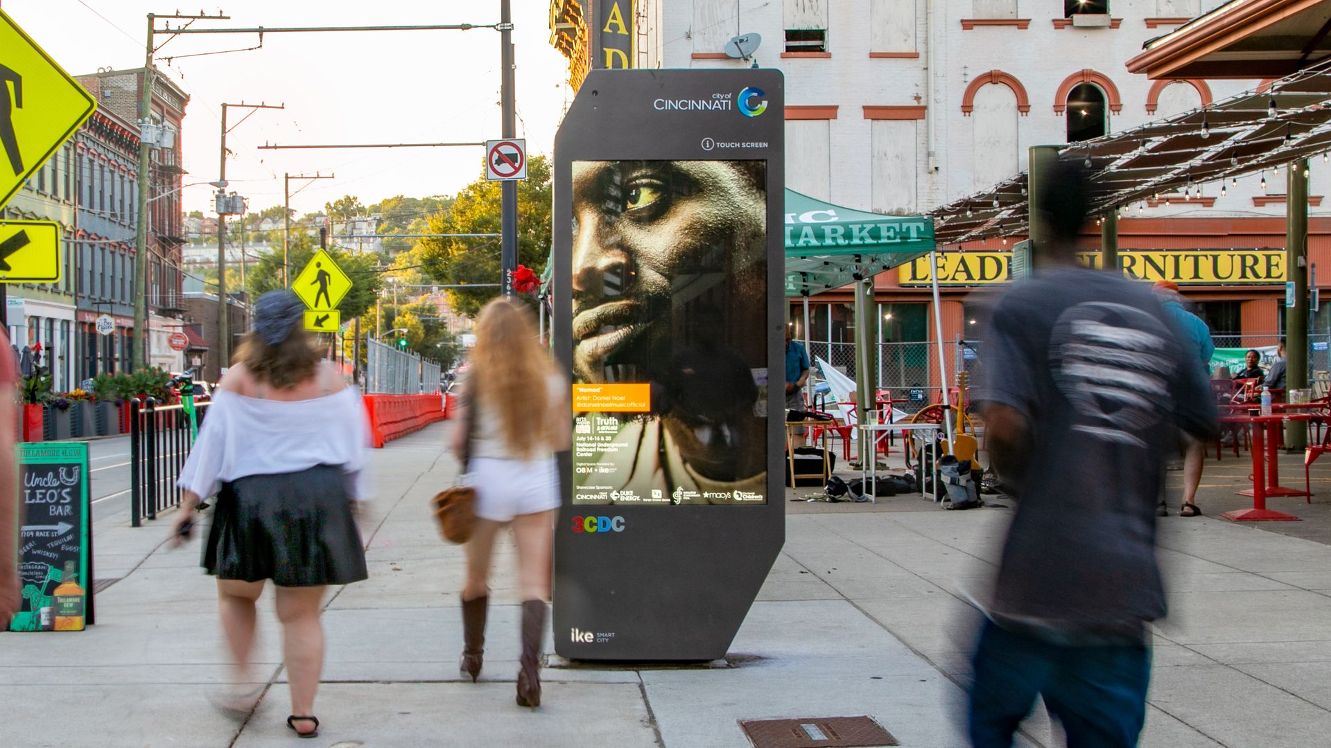 A digital kiosk on a sidewalk in Cincinnati as people walk by.