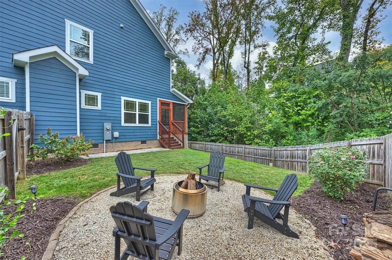 Backyard with blue house, green lawn, wooden fence, four black Adirondack chairs around a metal fire pit on gravel, surrounded by trees and plants under a partly cloudy sky.