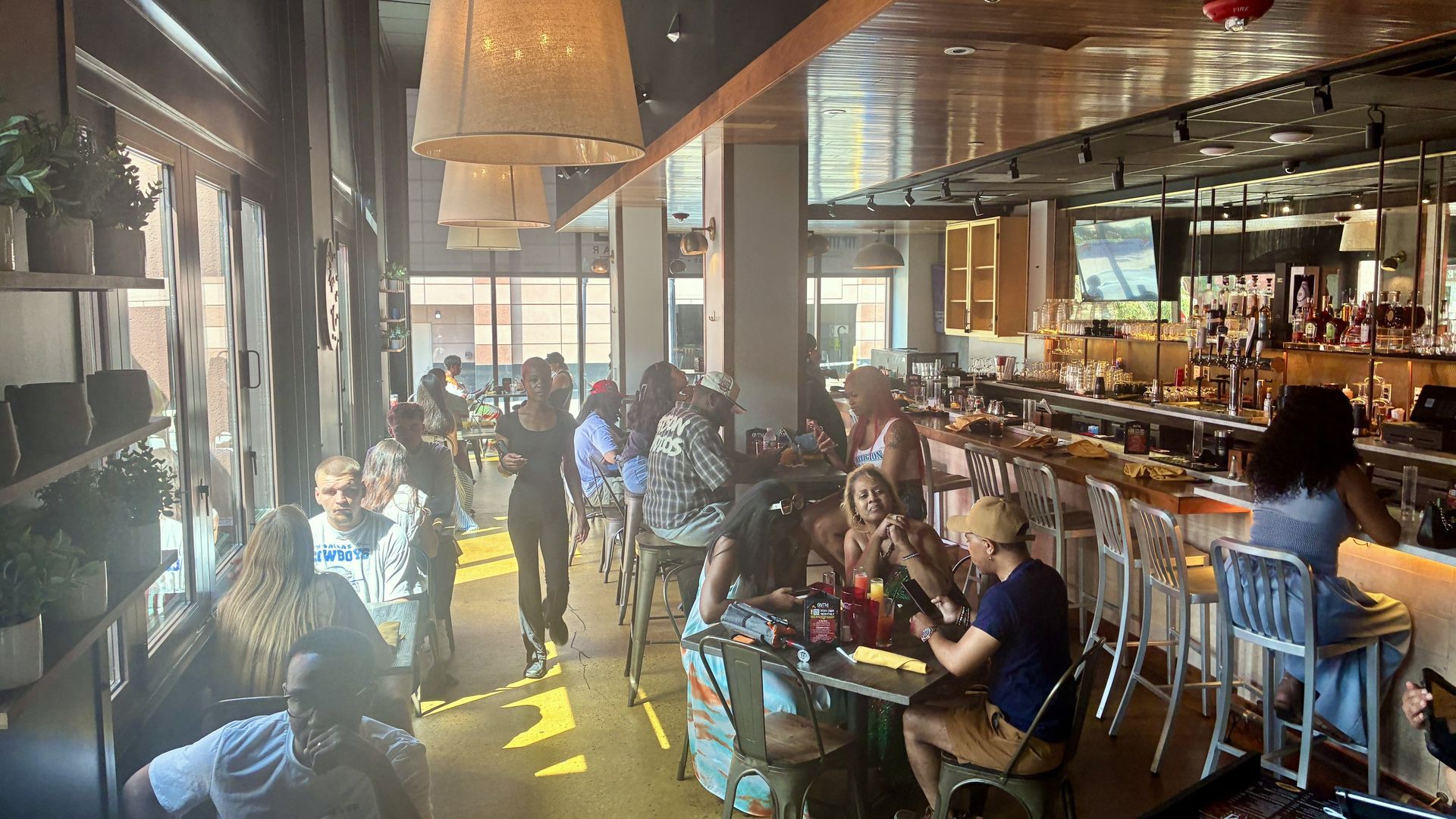 Busy restaurant interior with warm lighting, oversized hanging lamps, and a long bar on the right. Patrons sit at tables by windows on the left, chatting and using phones.
