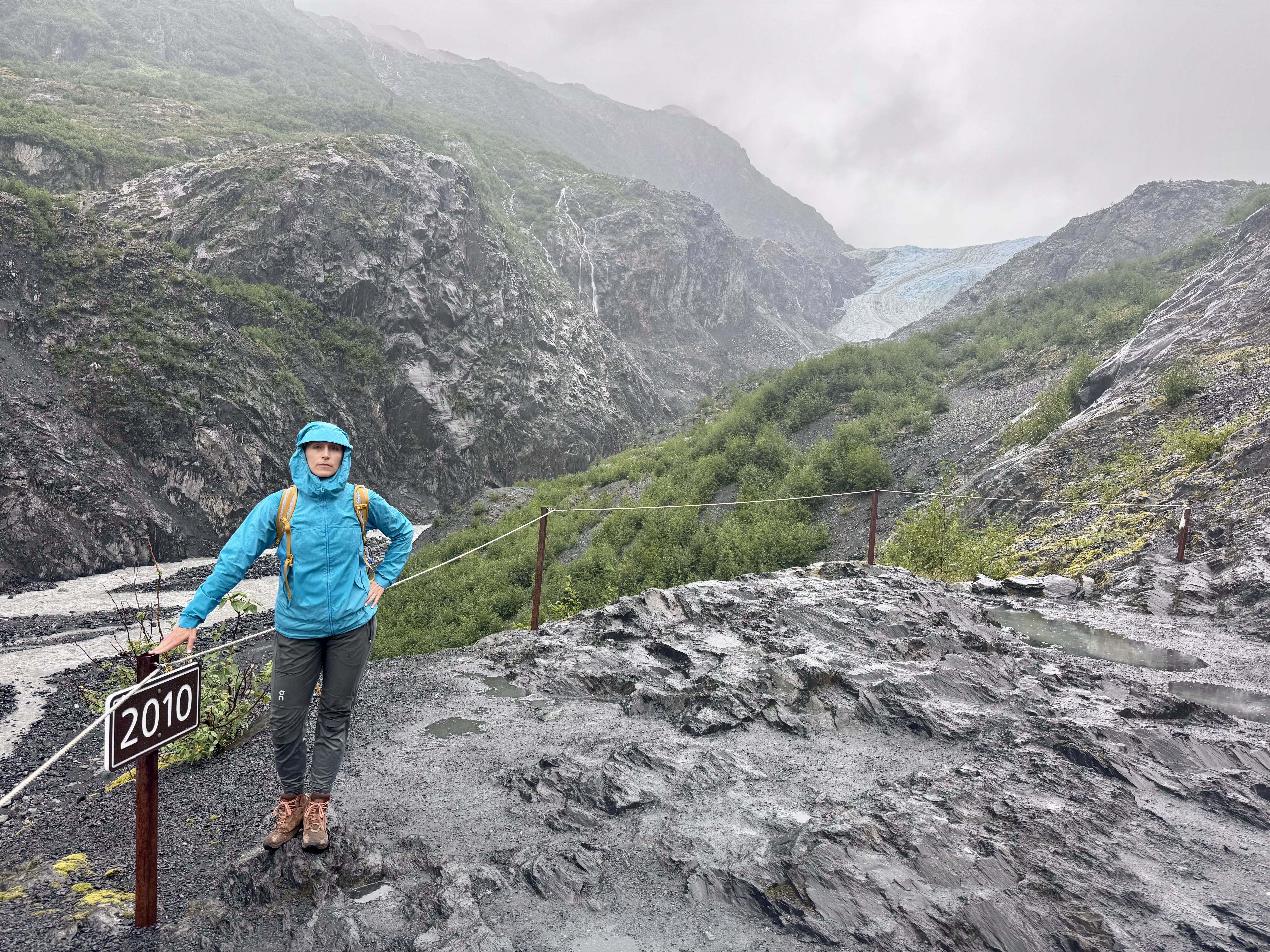 A hiker in a bright blue rain jacket and backpack stands on a wet rocky trail beside a rope barrier, in a canyon with dark jagged cliffs and a distant blue glacier under a cloudy sky.