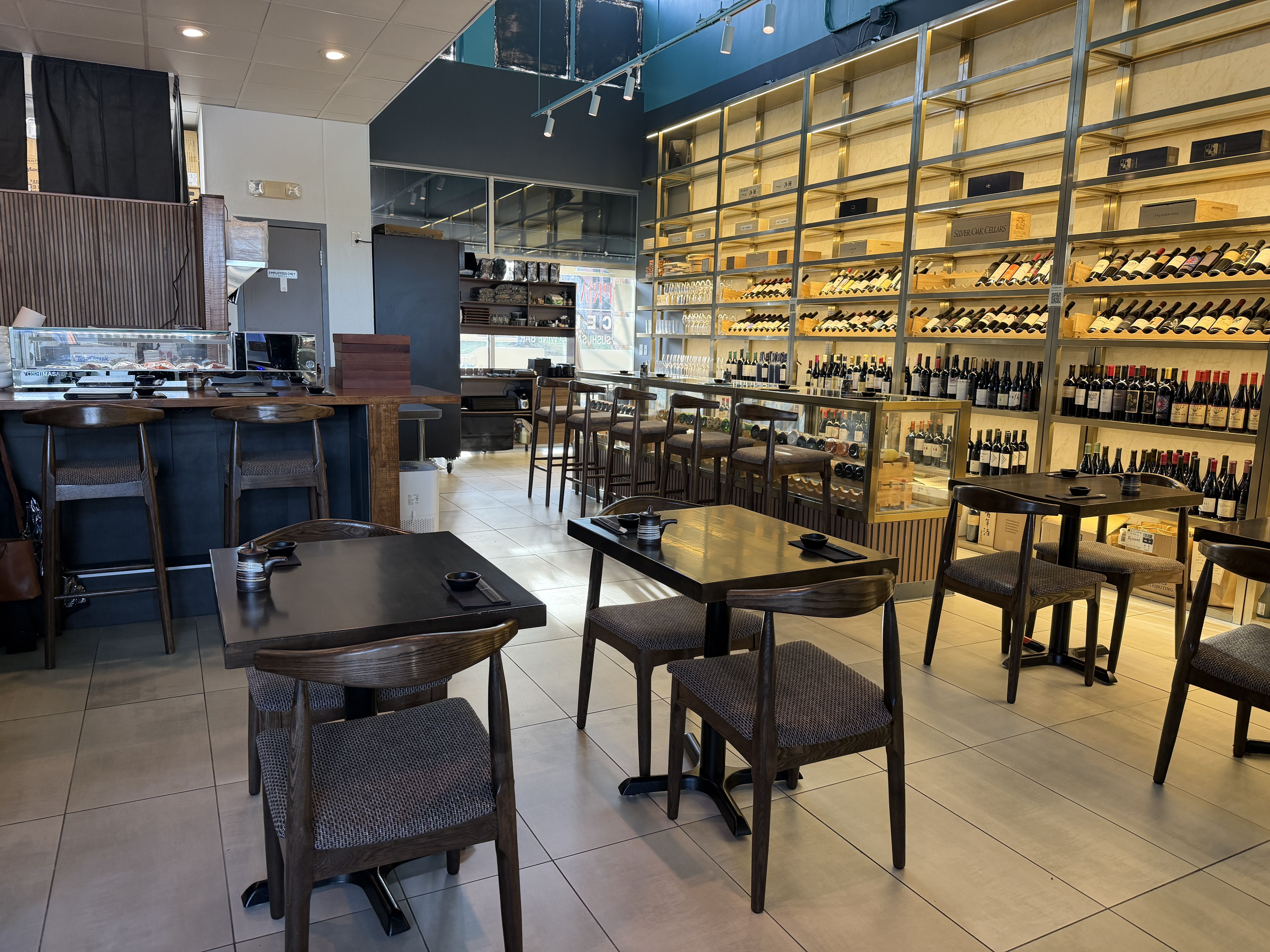 Modern wine bar interior with beige tile floor, dark wood tables and chairs, shelves filled with wine bottles and boxes, and a sushi preparation counter with stools.