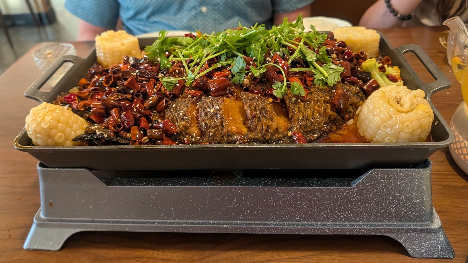 A grilled fish braised with sauce, chilis, corn and other items in a metal pan over a portable stove.