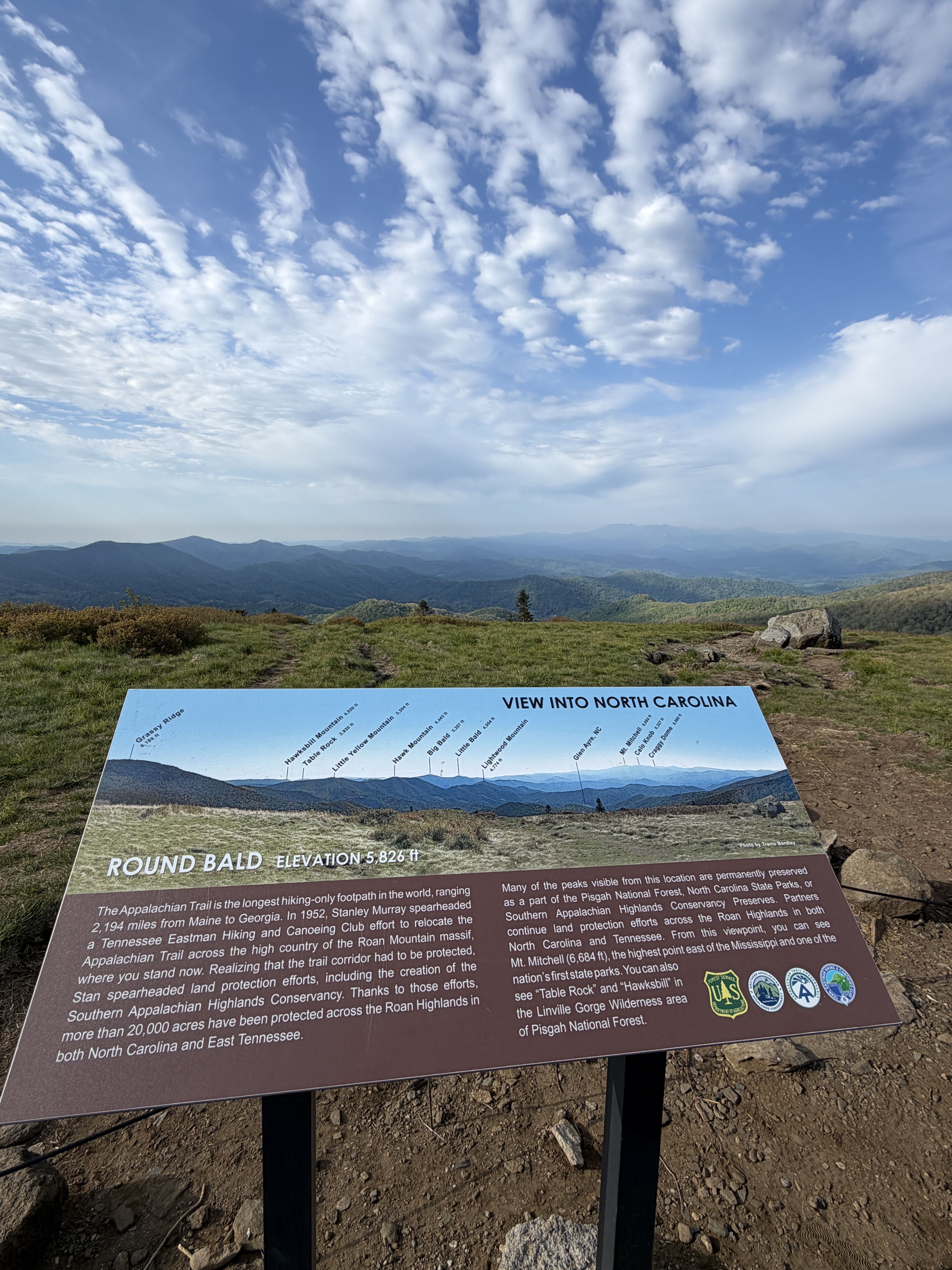 Panoramic mountain view under a bright blue sky with scattered clouds; in the foreground a large blue-brown sign reading "VIEW INTO NORTH CAROLINA" and "ROUND BALD ELEVATION 5,826 ft".