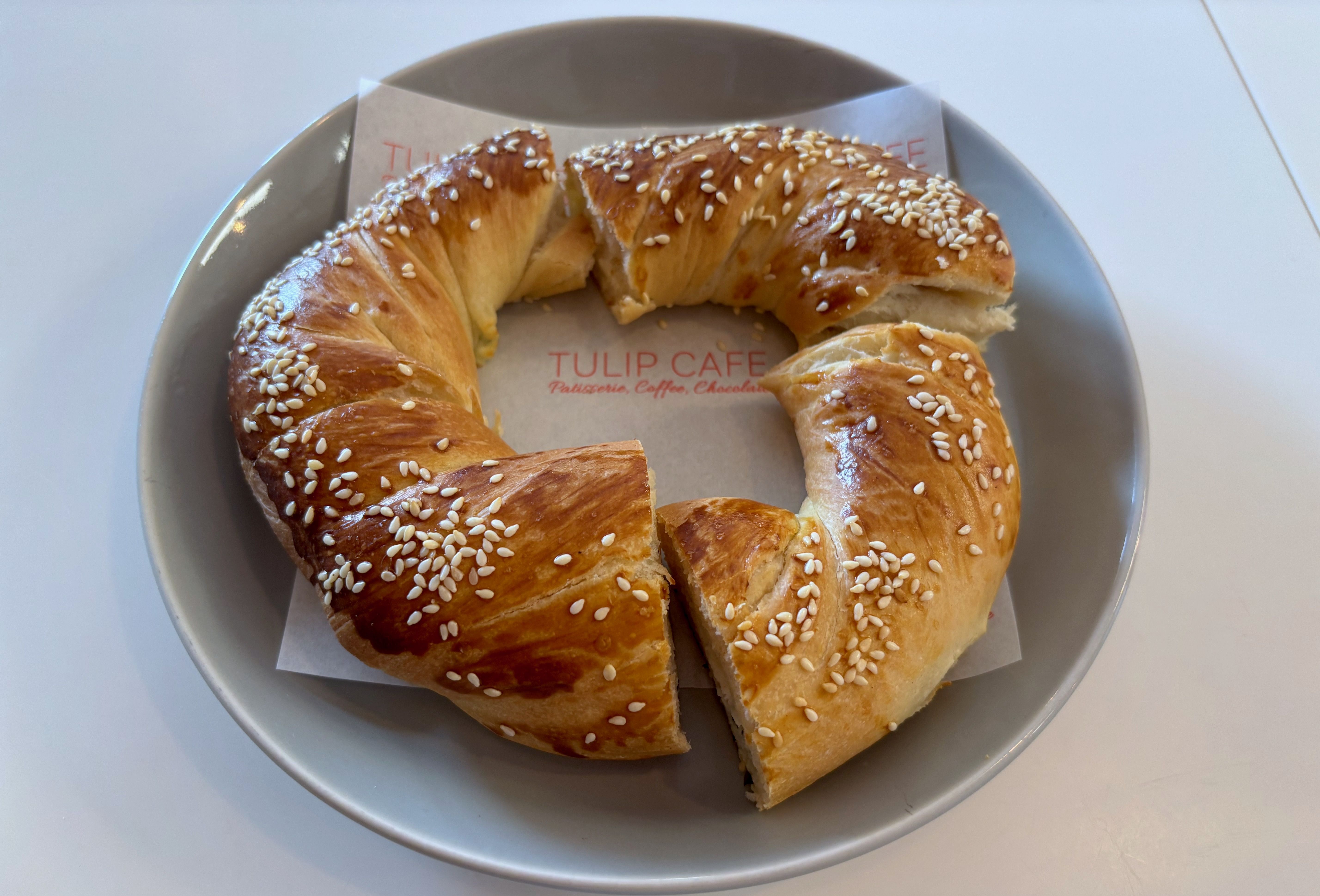 Golden-brown sesame bagel, sliced into quarters, rests on a gray plate with Tulip Café parchment; sesame seeds scatter across the shiny crust.