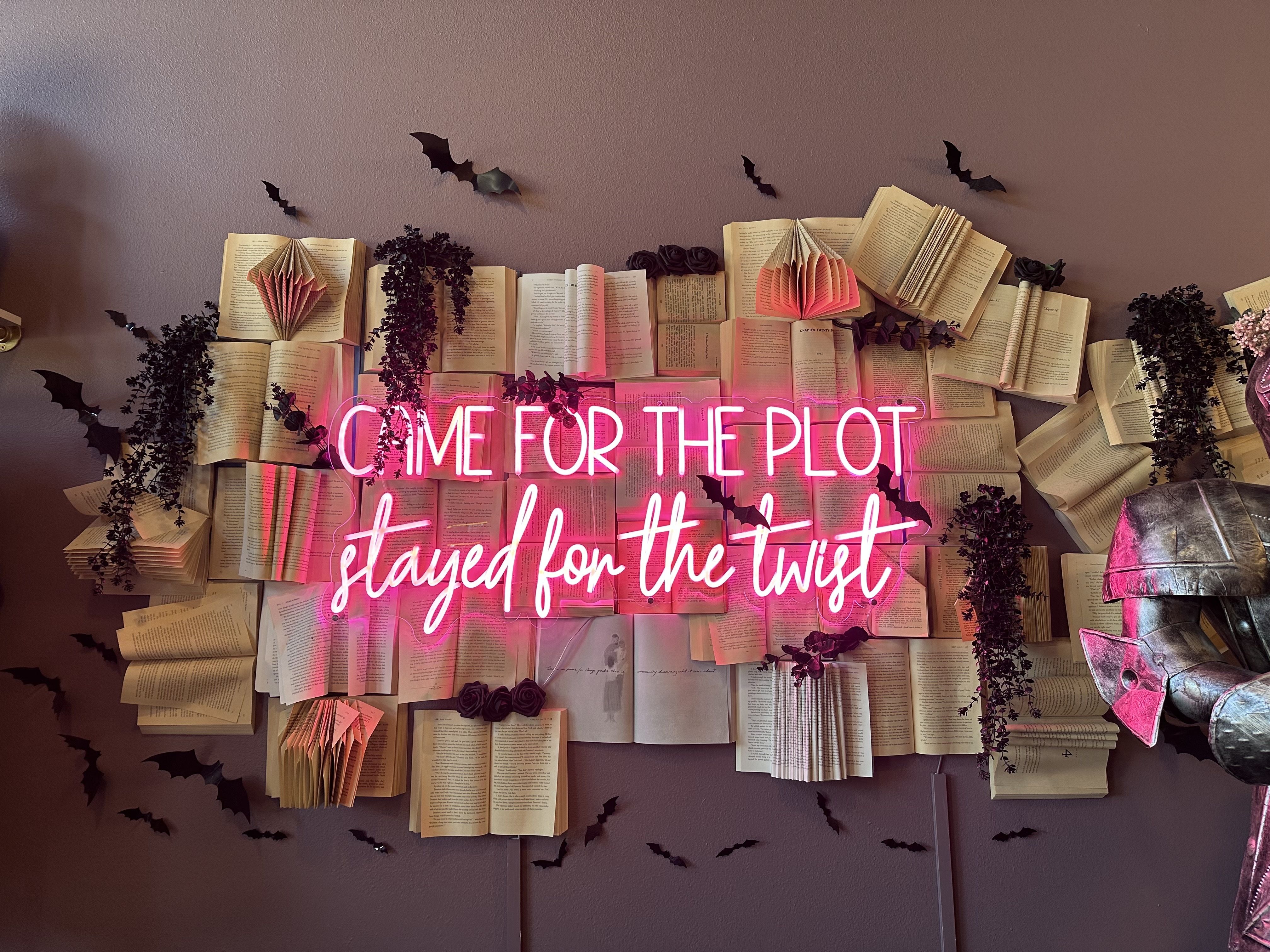 Wall decorated with open books, black paper bats, dark flowers, and hanging greenery; pink neon sign reads "COME FOR THE PLOT stayed for the twist"; partial metallic knight armor visible on right.