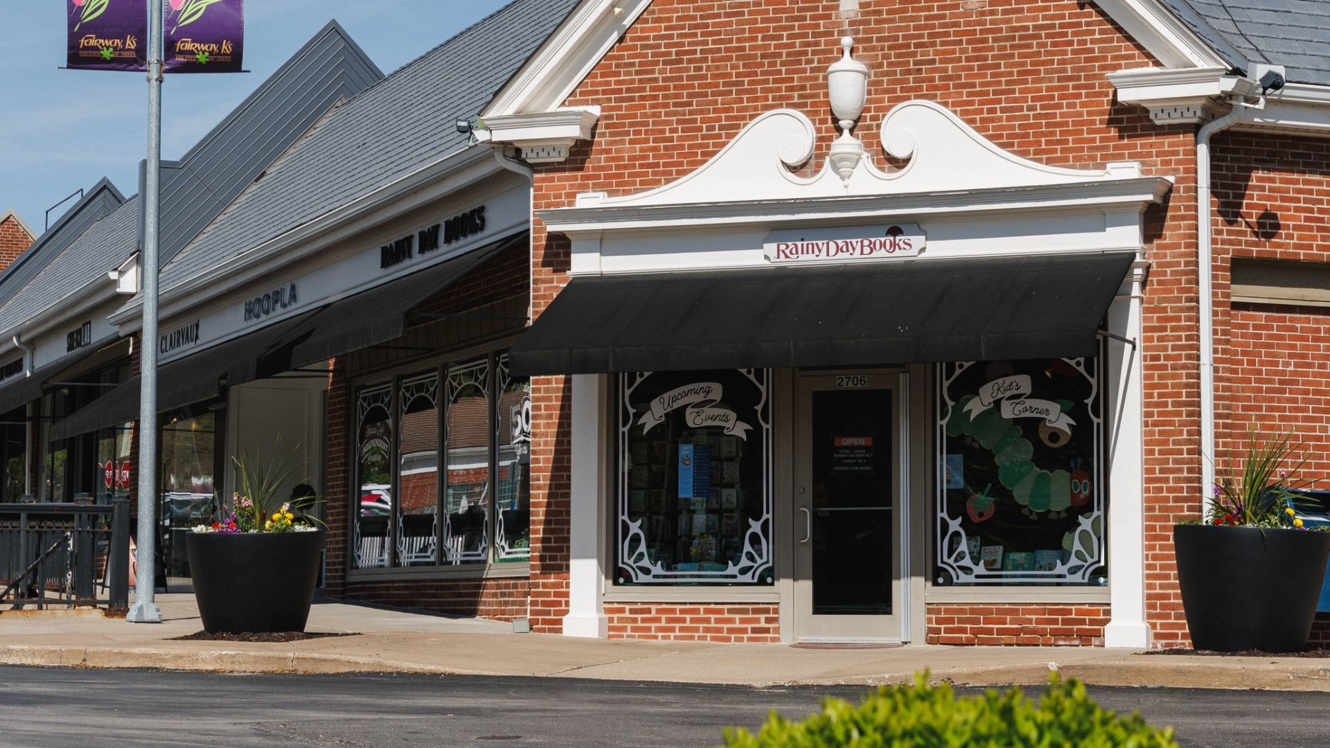 Exterior of a red brick bookstore named Rainy Day Books with black awnings and large windows displaying books and signs for "Upcoming Events" and "Kid's Corner."