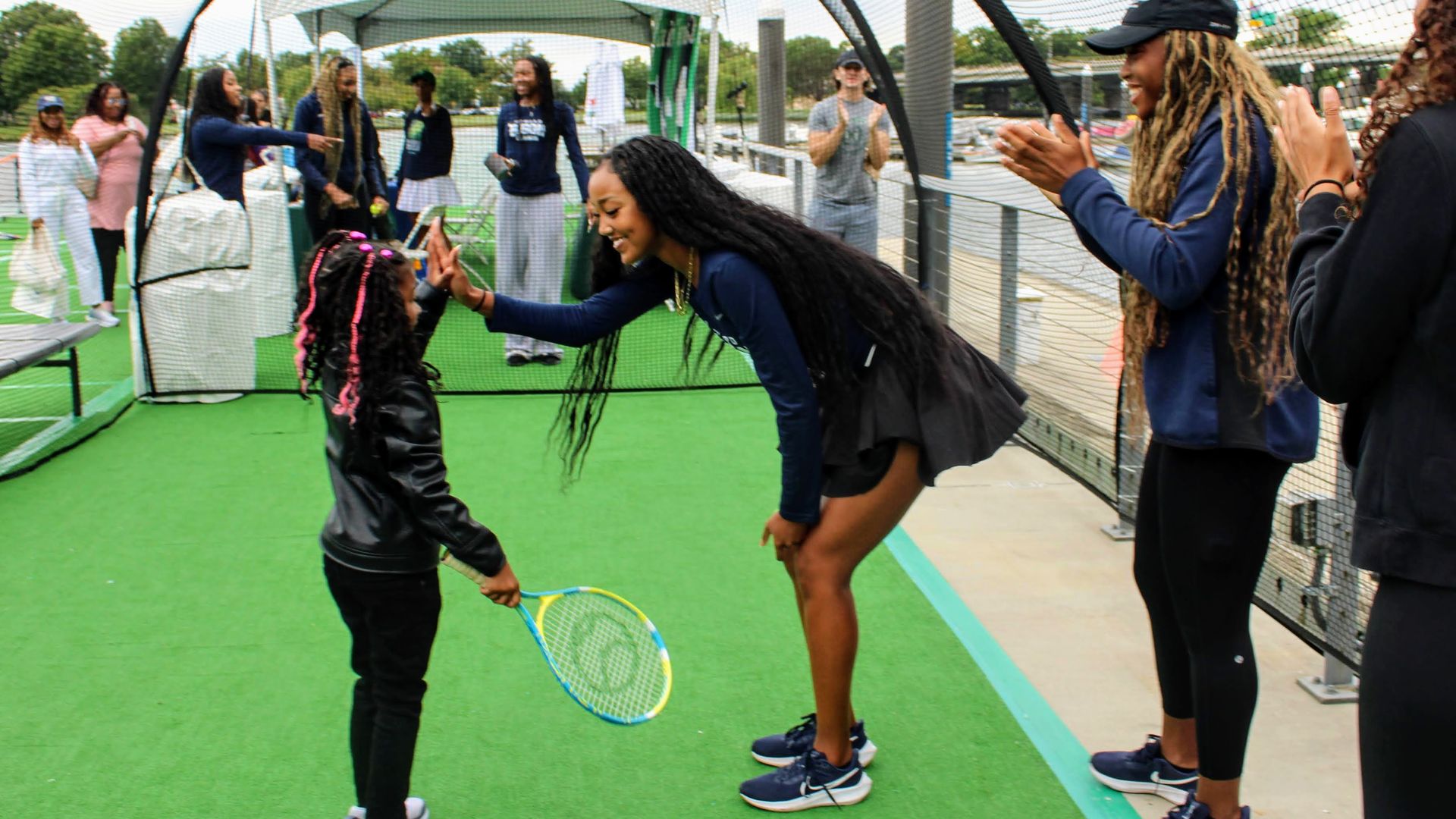 A young girl with pink braids holding a tennis racket high-fives a smiling woman with long hair in sportswear on green tennis court turf while others watch and clap nearby.