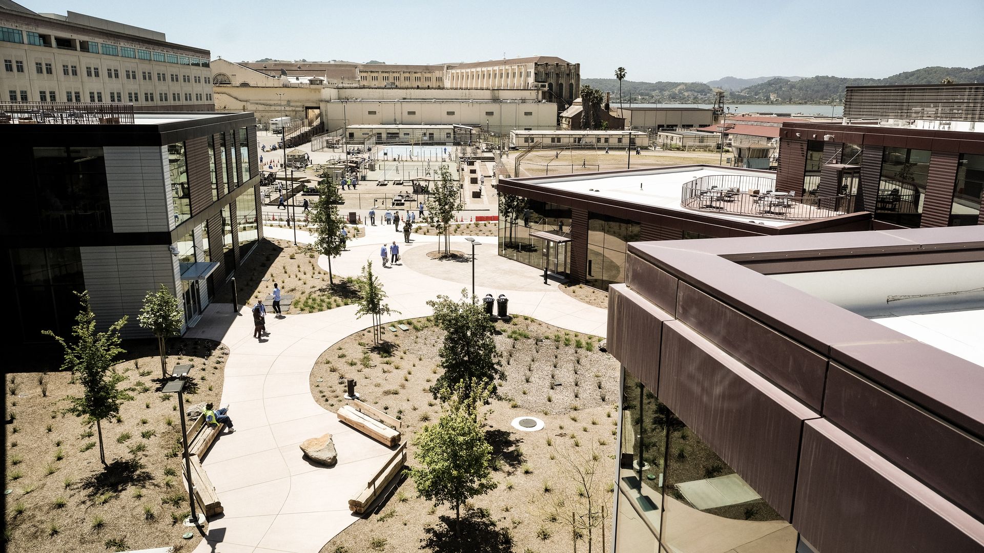 Wide campus courtyard flanked by modern glass buildings; a central plaza with trees, benches, and pedestrians; distant beige industrial building, hills and water under a clear blue sky.