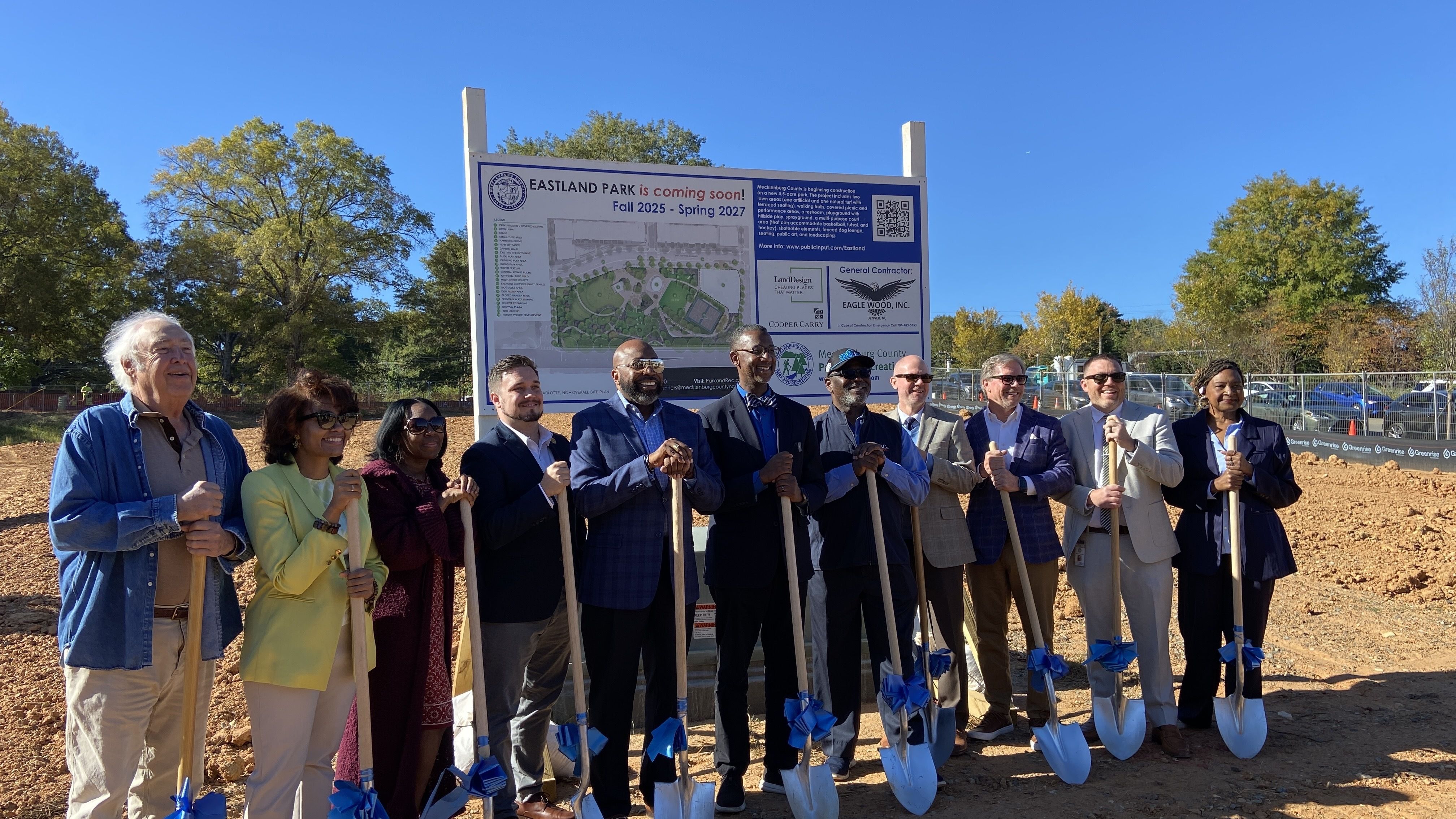 Group of eleven people holding shovels with blue bows at a groundbreaking event for Eastland Park under clear blue sky with a project sign behind them.