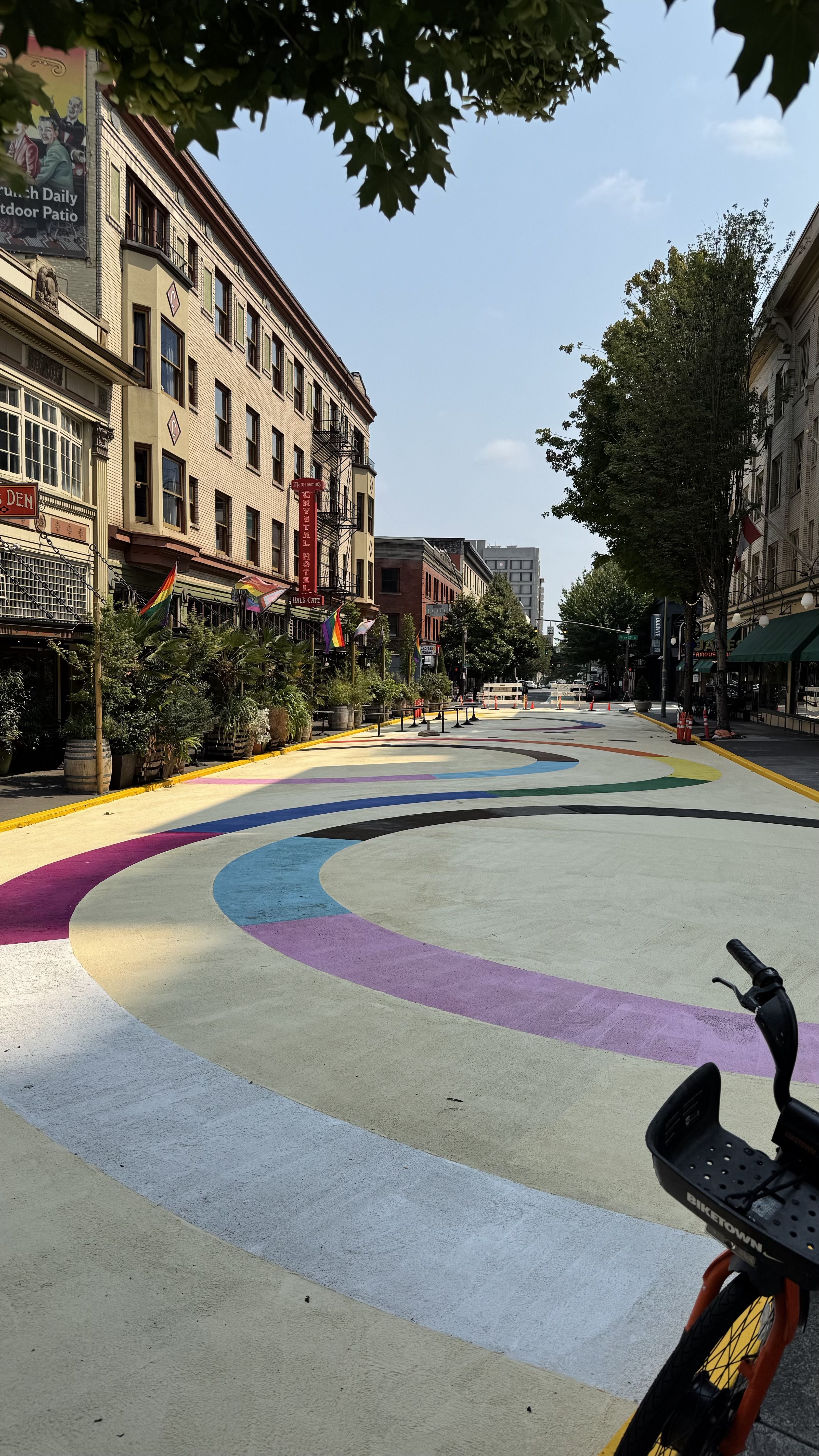 Sunlit city street with brick buildings on the left and trees on the right. Rainbow curved pavement winds along the street; rainbow flags line the sidewalk. A bicycle in the foreground.