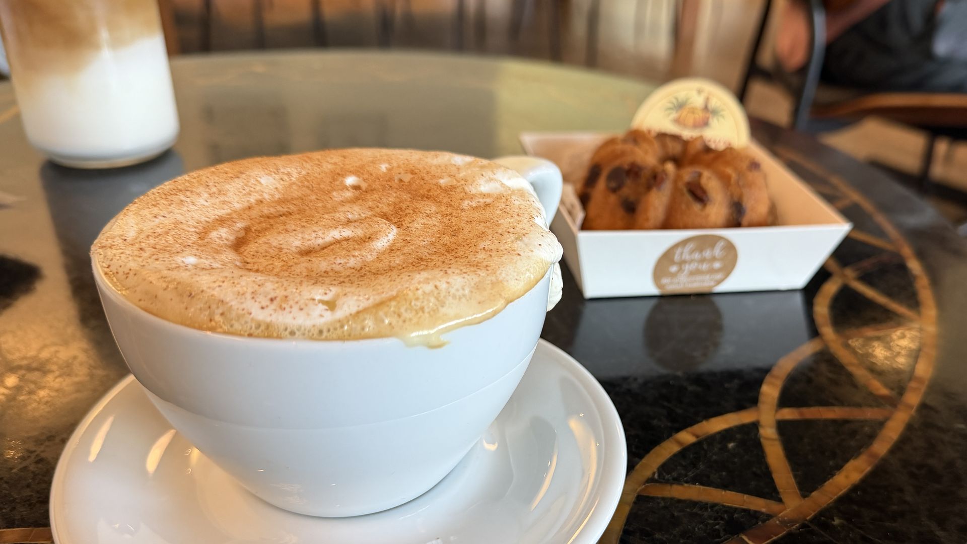 White cup of frothy cappuccino with cinnamon dusting on a saucer, in the foreground; a pastry in a white box sits in the background on a café table.