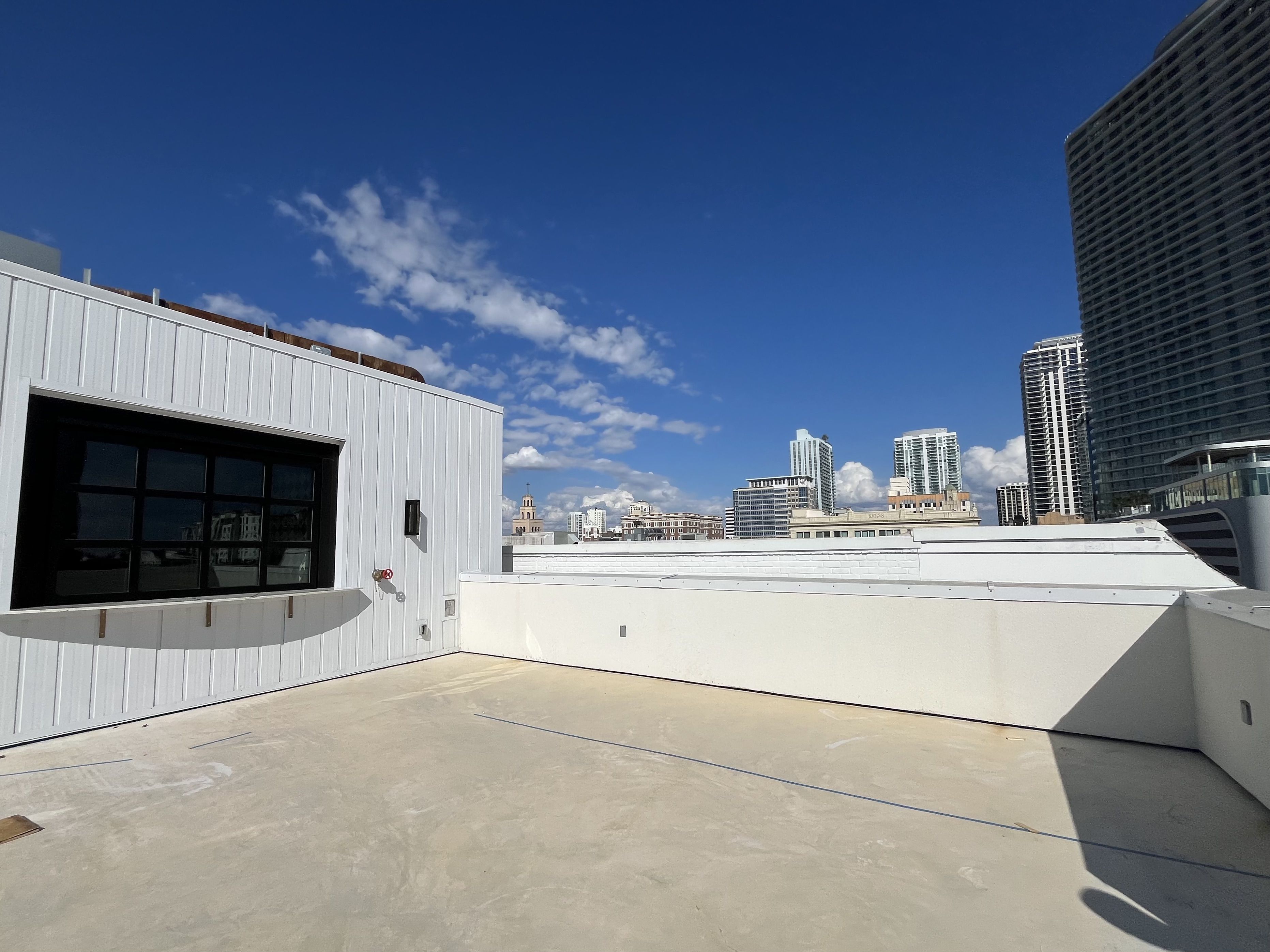 Bright rooftop terrace with white walls and beige floor under clear blue sky, city skyline with tall buildings in the distance, and a white building with black window on left.