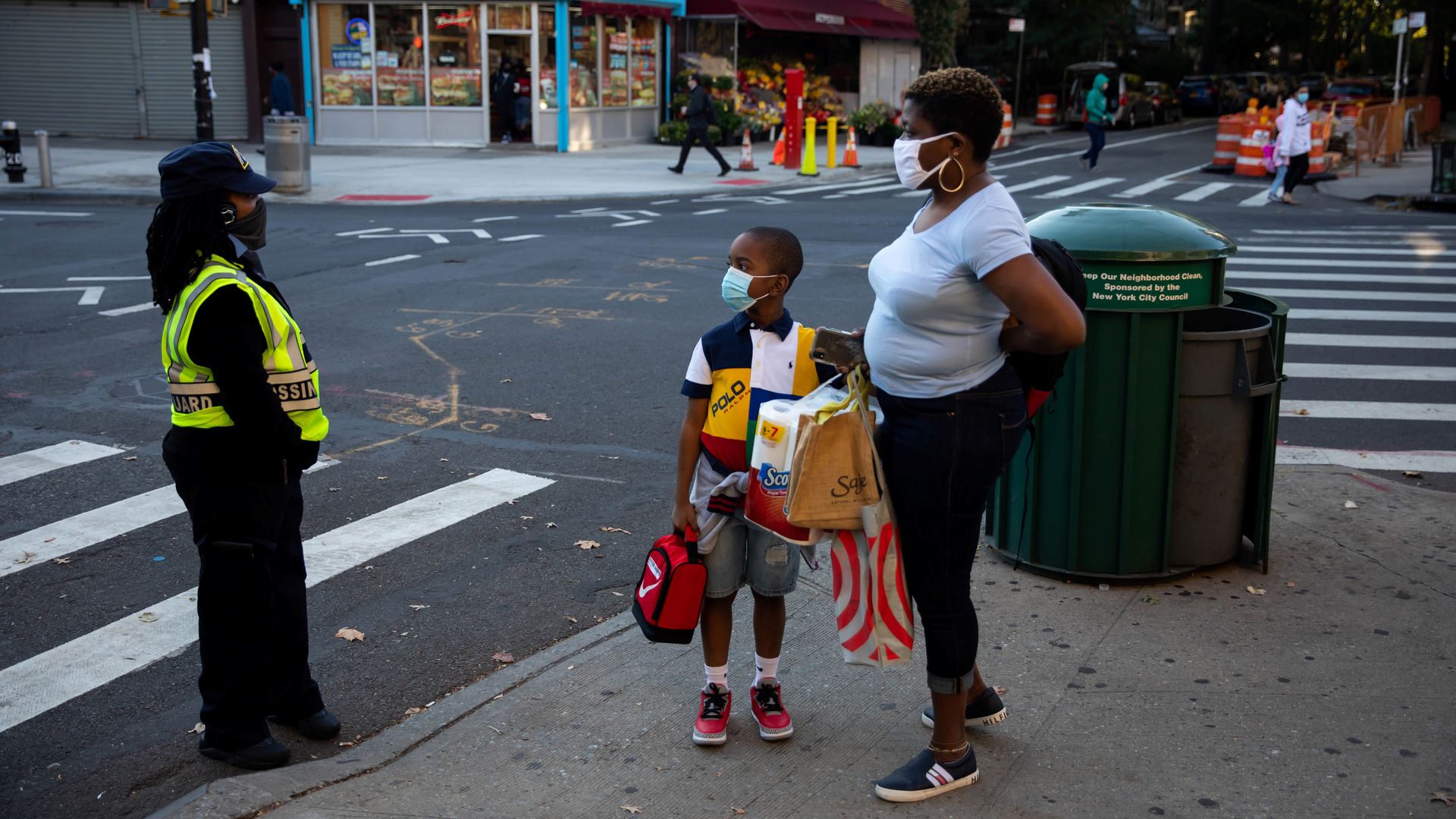 A student stands by his mother while holding a lunchbox and his backpack, while a crossing guard in a bright yellow vest stands near them and the street