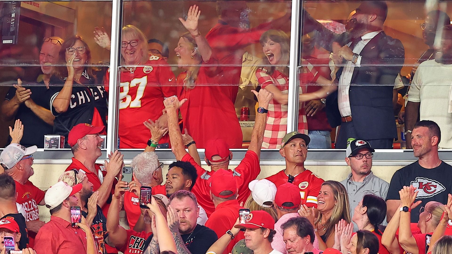 Taylor Swift wearing a gingham red dress cheers along with Jason and Donna Kelce in a box with Chiefs fans in front all dressed in red. 