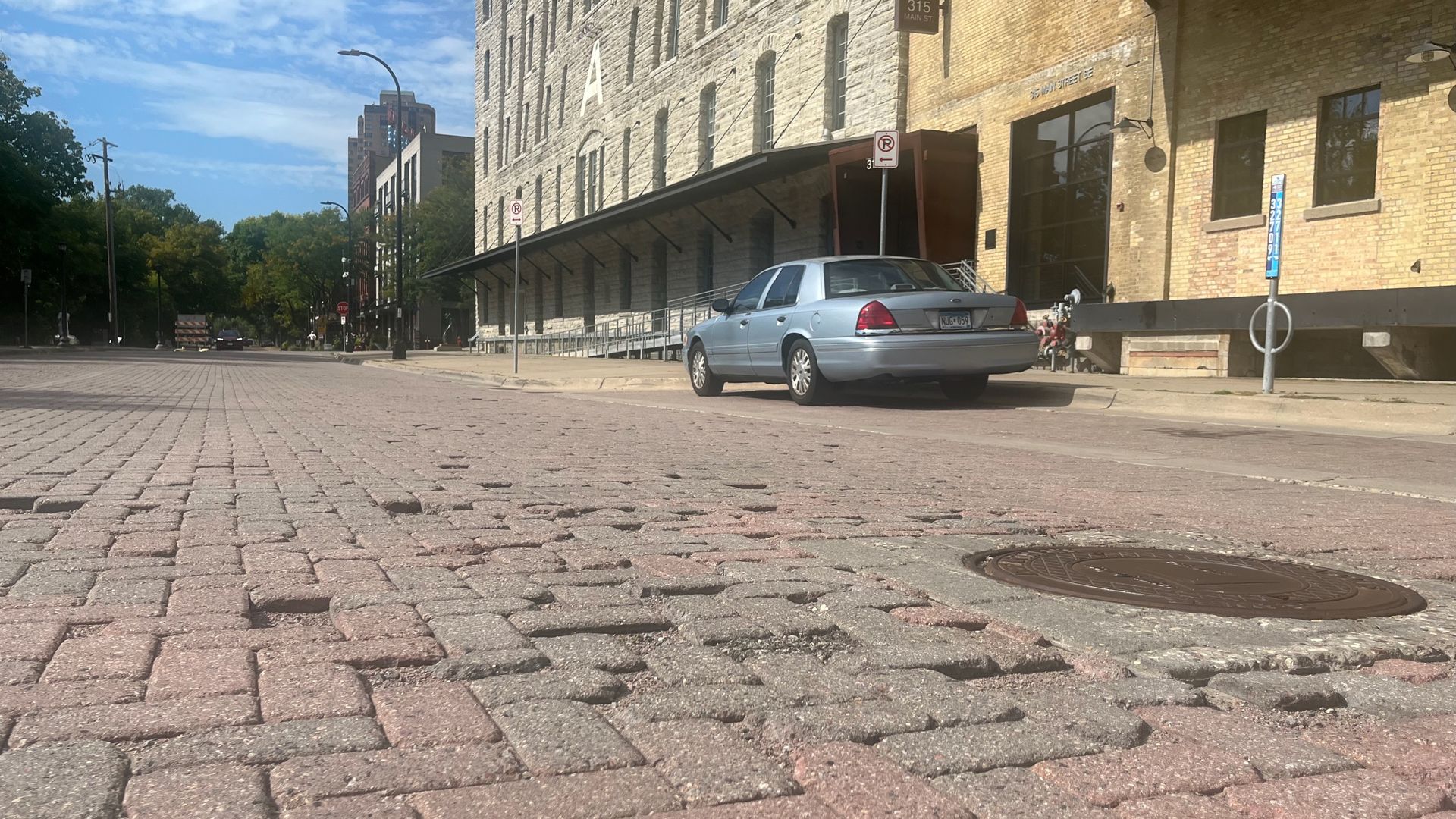 Low-angle view of a brick-paved street with cracks and uneven bricks, a silver car parked by a beige brick building with tall windows under a blue sky with clouds.