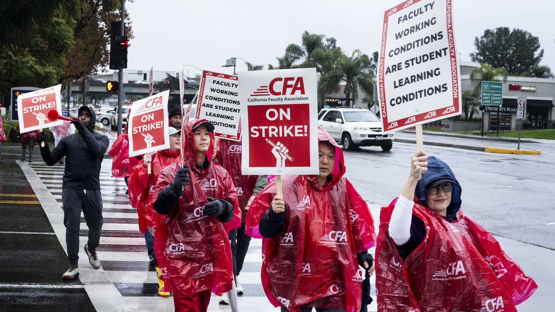 Faculty members wearing red rain ponchos walk across the street on a college campus holding signs during a strike.