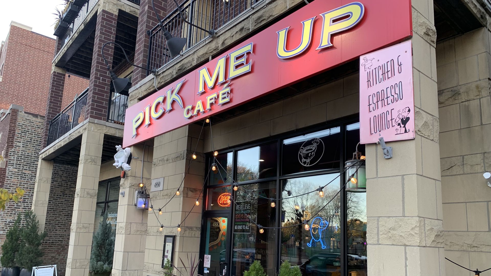 Cafe exterior with a red sign reading "PICK ME UP CAFÉ" in yellow letters, string lights, potted plants, a pink sign saying "KITCHEN & ESPRESSO LOUNGE," and an open neon sign.