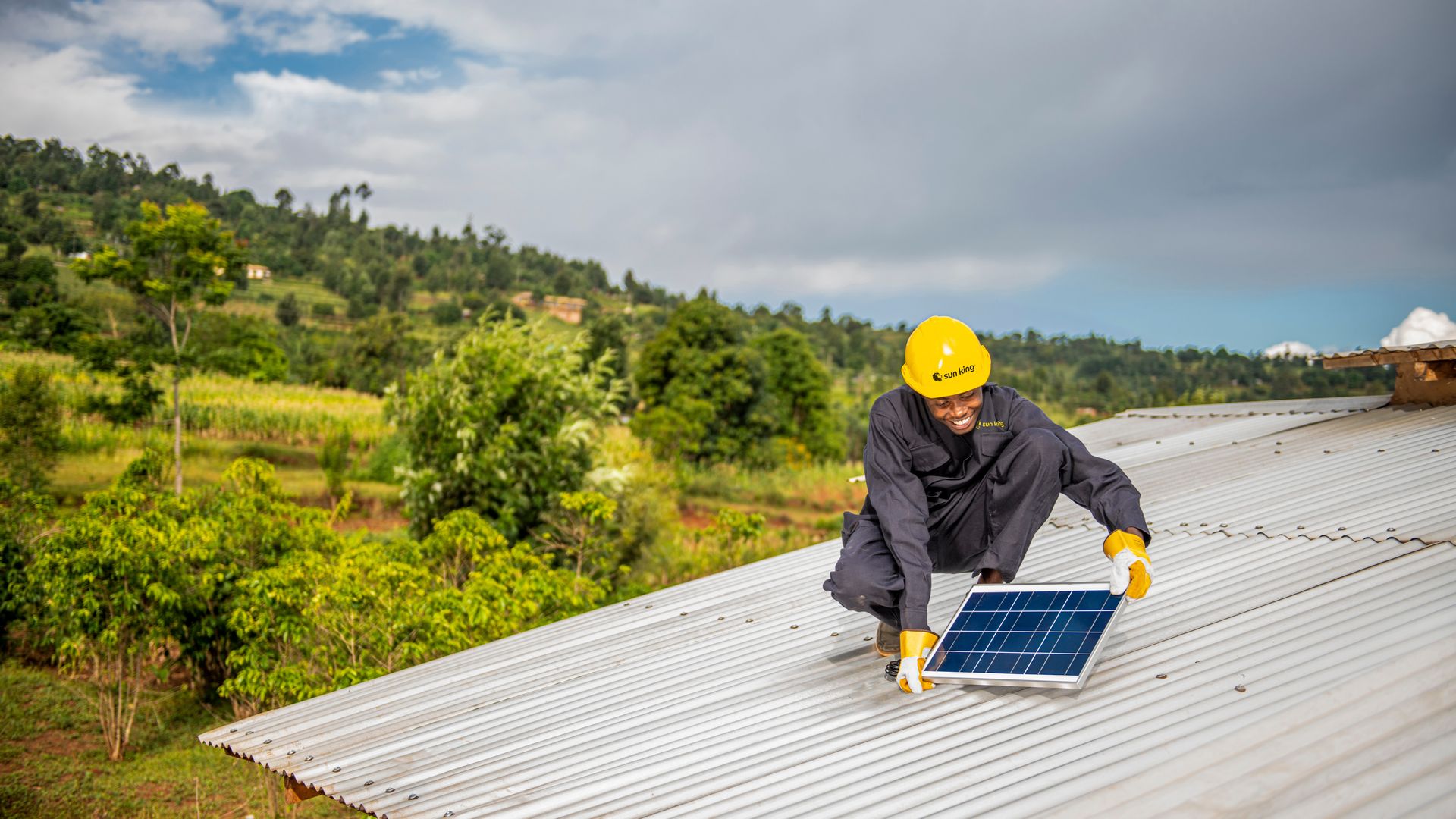 Man in black coveralls and yellow helmet installing a solar panel on a silver metal roof with green trees and hills in the background under a partly cloudy sky.