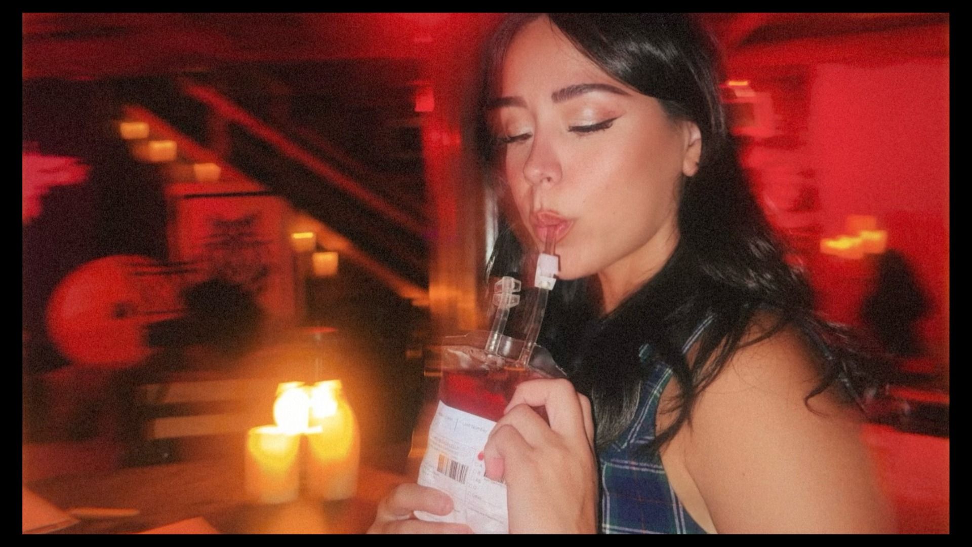 Young woman with dark hair and winged eyeliner in a red-lit bar, wearing a plaid top, closes her eyes while sipping from a bottle. Warm candles glow in the foreground; blurred background.
