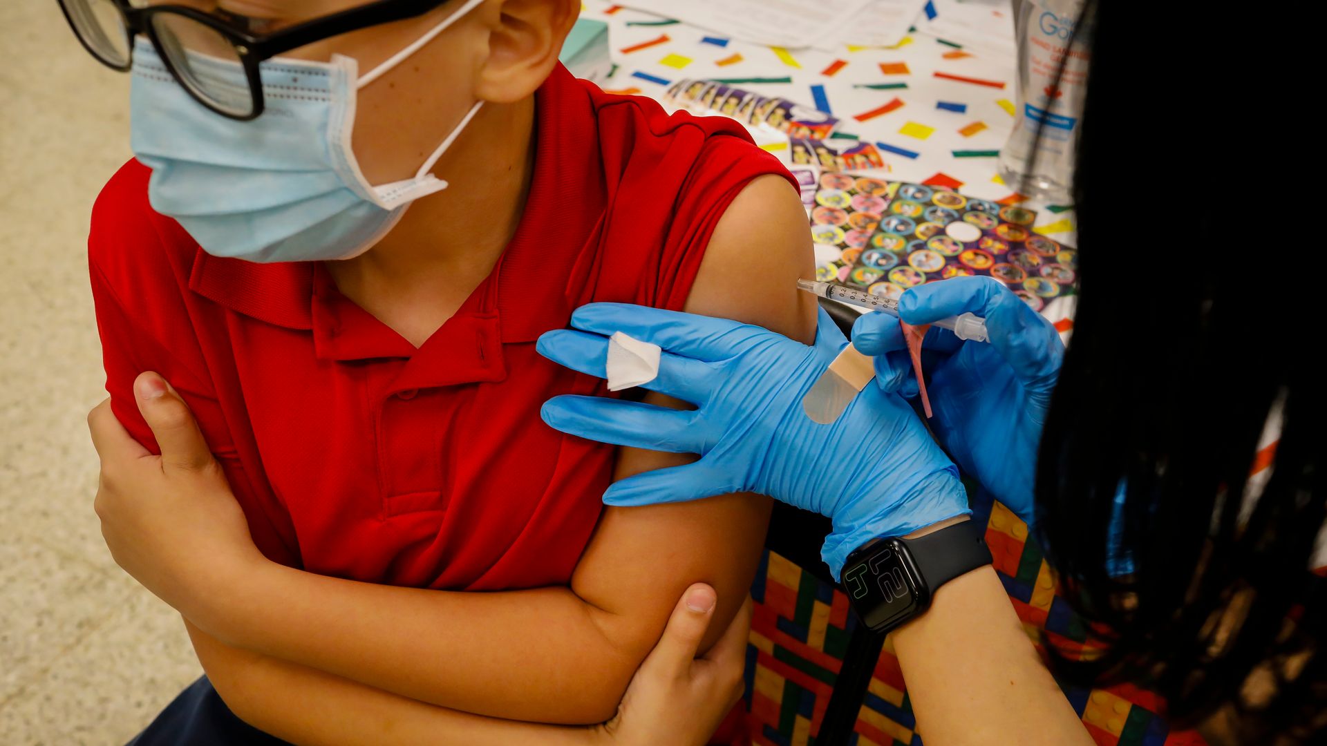 Photo of a child in red with their sleeve rolled up as a nurse injects a COVID shot into their left arm