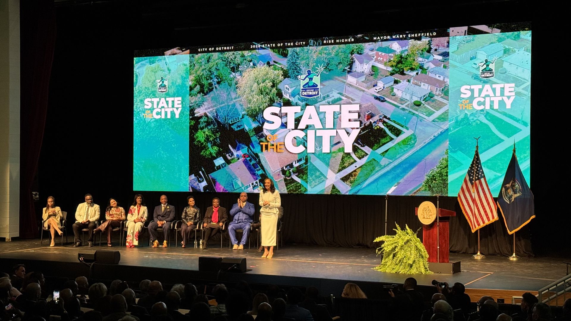 A stage with a screen saying "State of the City" in the background. Mayor is in the foreground standing and speaking, with city council members behind