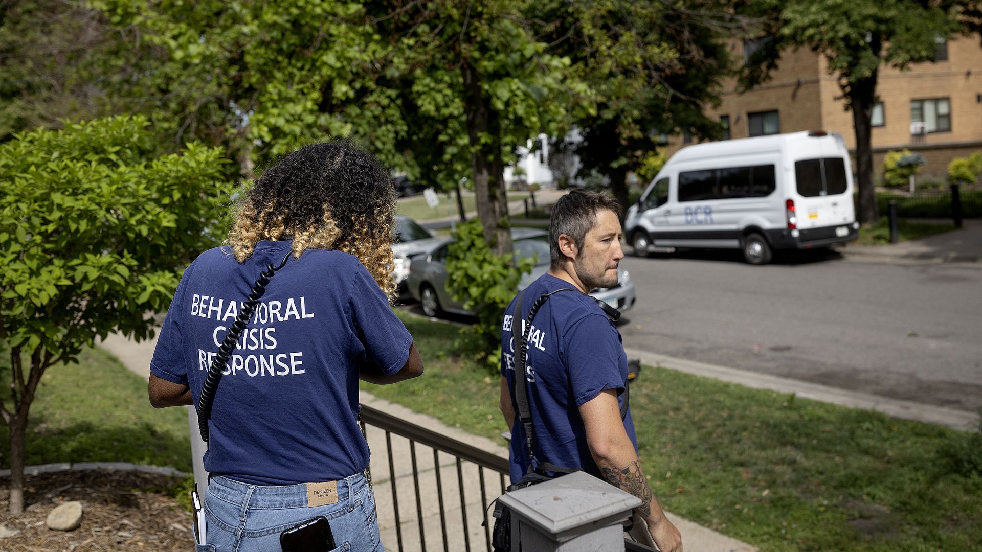 Two crisis responders in navy shirts bearing 'BEHAVIORAL CRISIS RESPONSE' walk along a sidewalk amid greenery, with a white van and brick apartment building in the background.
