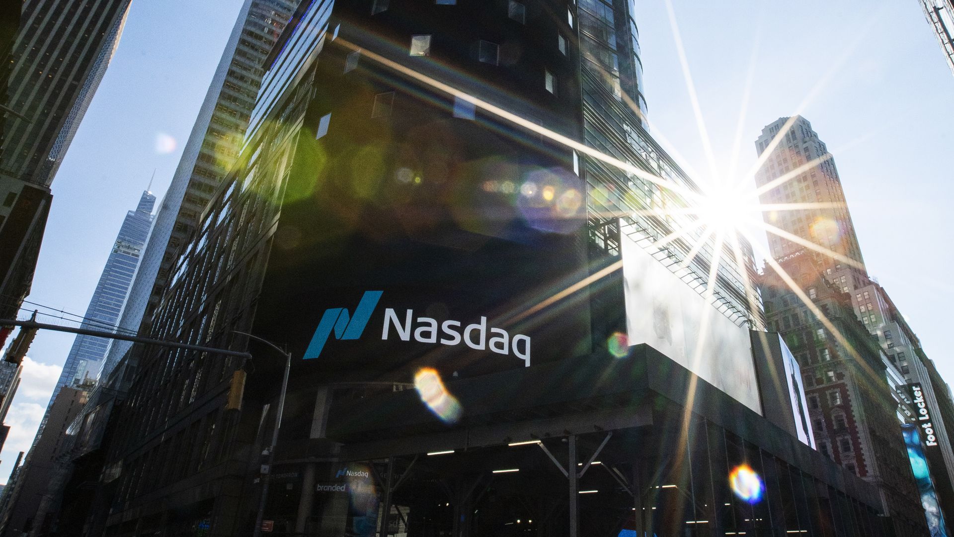 Urban cityscape with tall glass skyscrapers; a rounded dark building dominates the center. Bright sunburst lens flare on the right, blue sky, and a Nasdaq sign on the storefront.