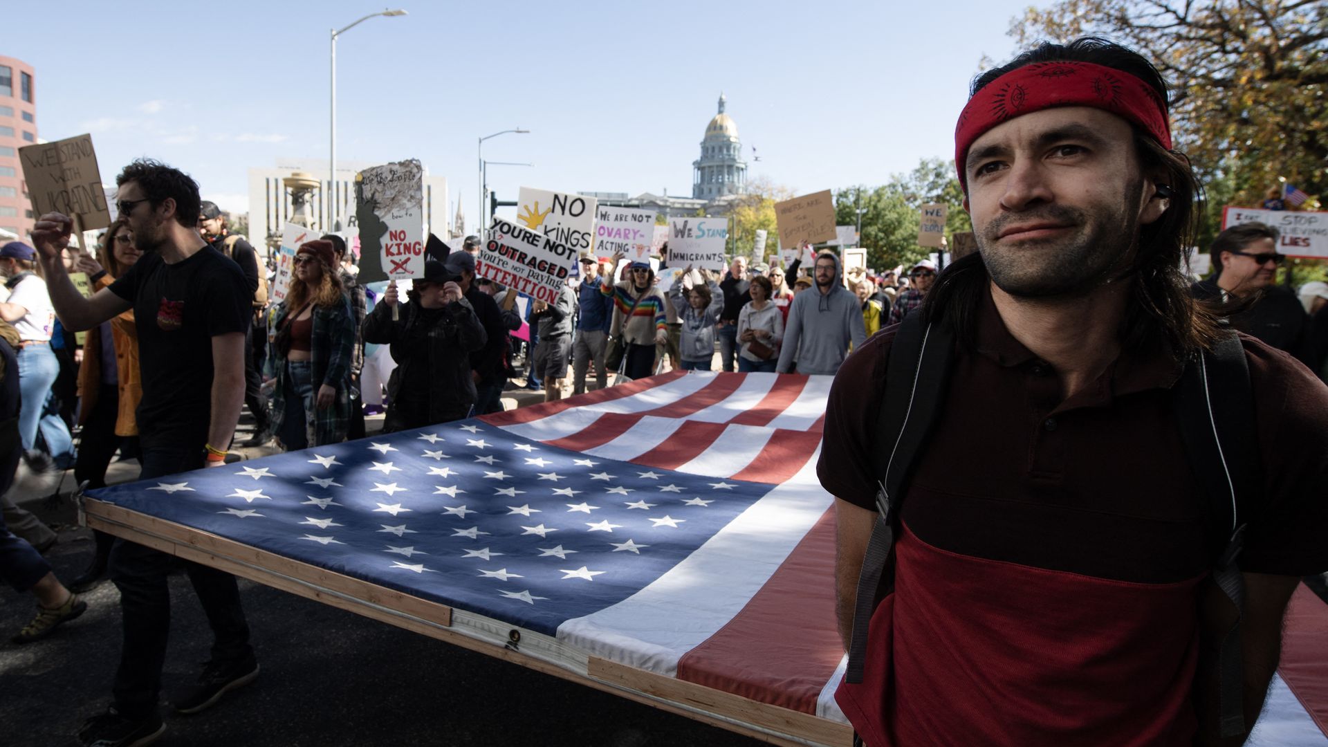 Protesters march down a street, carrying a large American flag banner; signs rise behind them as a man with a red bandana smiles in the foreground.
