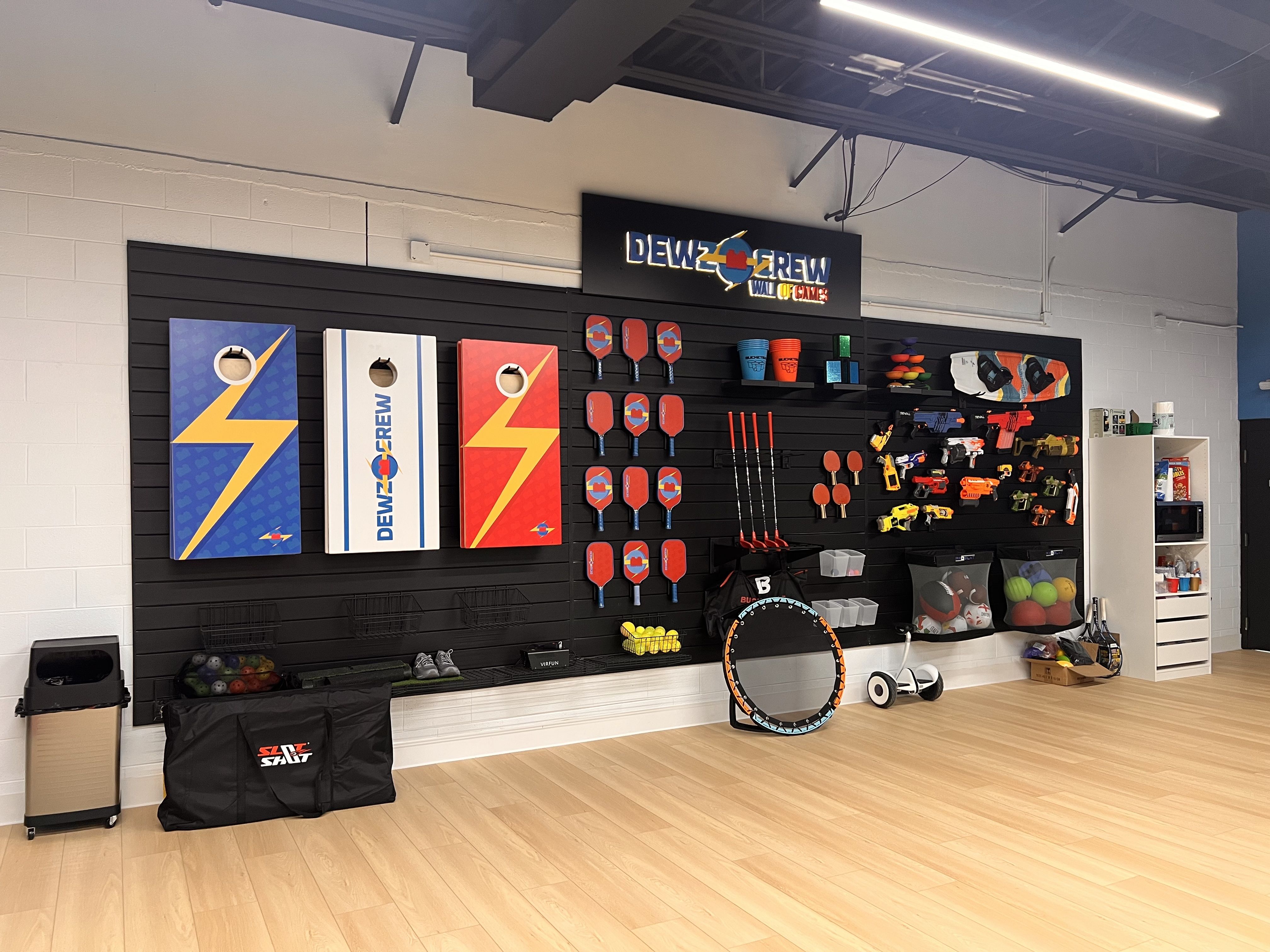 Indoor game store wall display with a "DEWZ CREW" sign; colorful paddles, rackets, and gear on a black pegboard, plus shelves of toys on a light wood floor under bright lights.