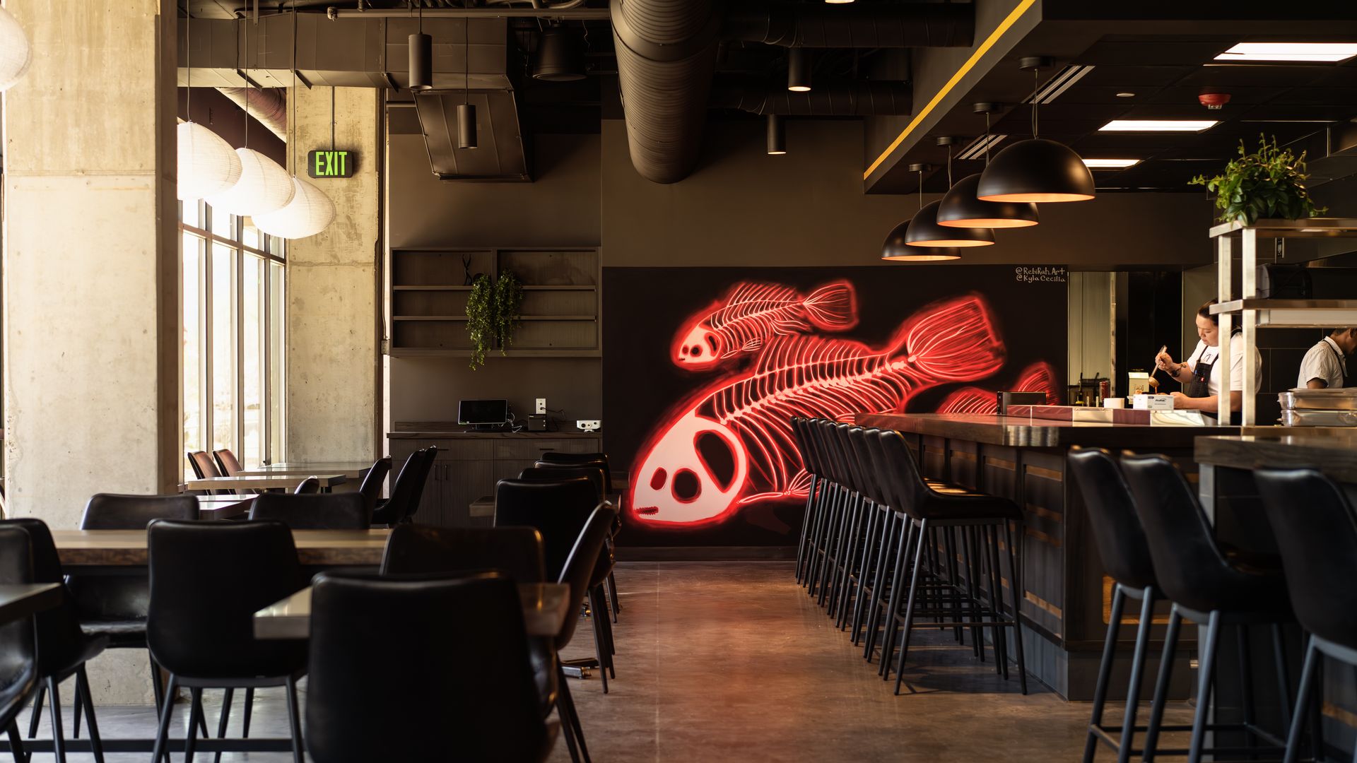 Industrial restaurant interior with concrete columns, black furniture, and a red neon skeleton fish mural on a dark wall behind the open kitchen; black bar stools line a wooden counter.